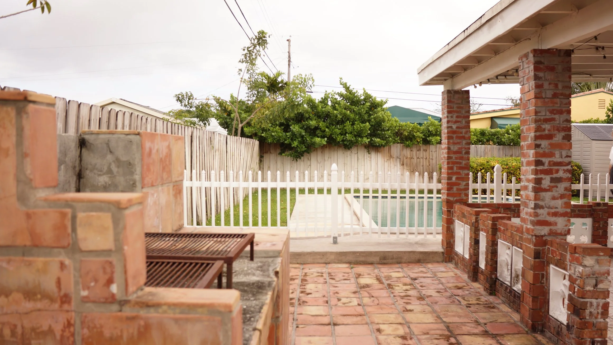 A backyard patio with brick built-in grill, tiled floor, and a swimming pool enclosed by a white picket fence. There is a wooden bench and a privacy fence with trees and neighboring houses in the background.