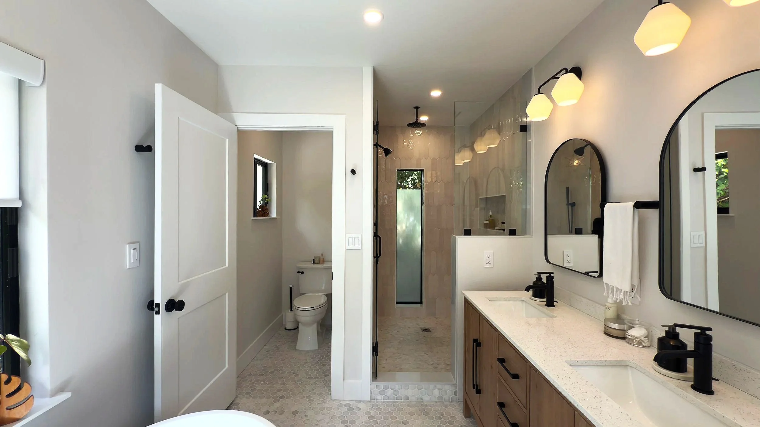 Modern bathroom with double vanity, large mirrors, black fixtures, wall-mounted lights, walk-in shower with glass door, toilet, small window, and neutral color palette.