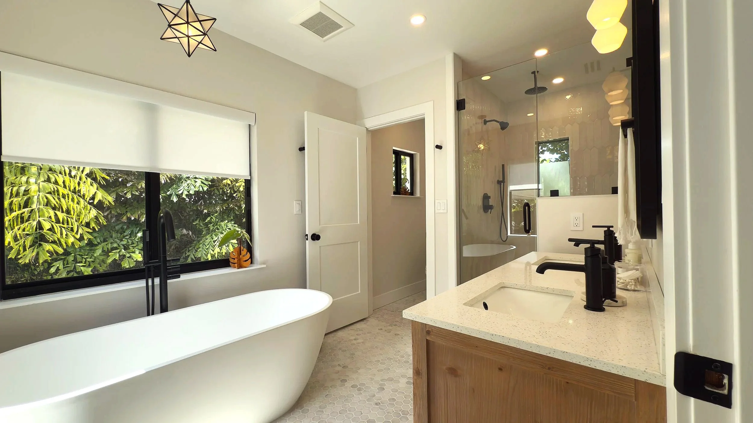 Modern bathroom with a freestanding bathtub near a large window with greenery outside, a glass-enclosed shower, a wooden vanity with black fixtures, and star-shaped ceiling light fixtures.