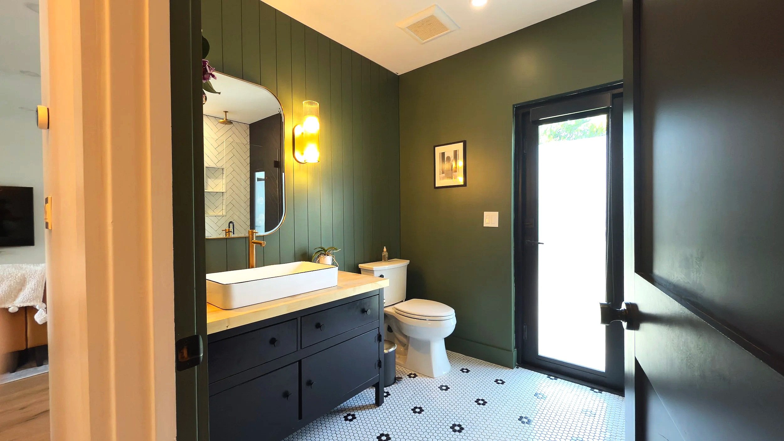 Modern bathroom with dark green walls, a white vessel sink with gold fixtures, a mirror, a framed picture, and a black vanity. There is a white toilet, a small plant, and a light-colored countertop. A black-framed glass door leads outside, and black 
