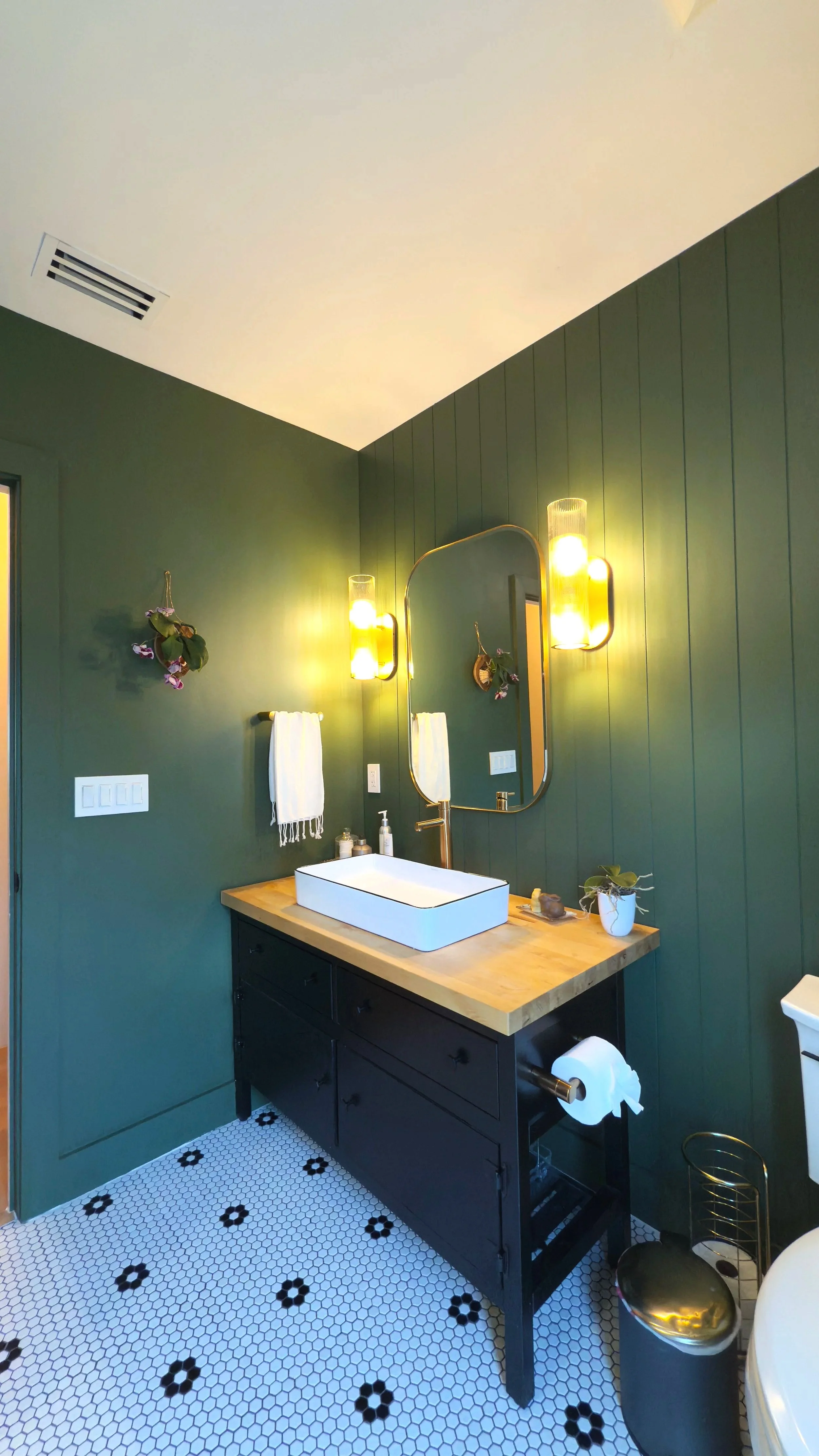 Modern bathroom with green paneled walls, black vanity with wooden countertop, white rectangular vessel sink, oval mirror, yellow wall sconces, white towels, plants, and black and white hexagon tiled floor