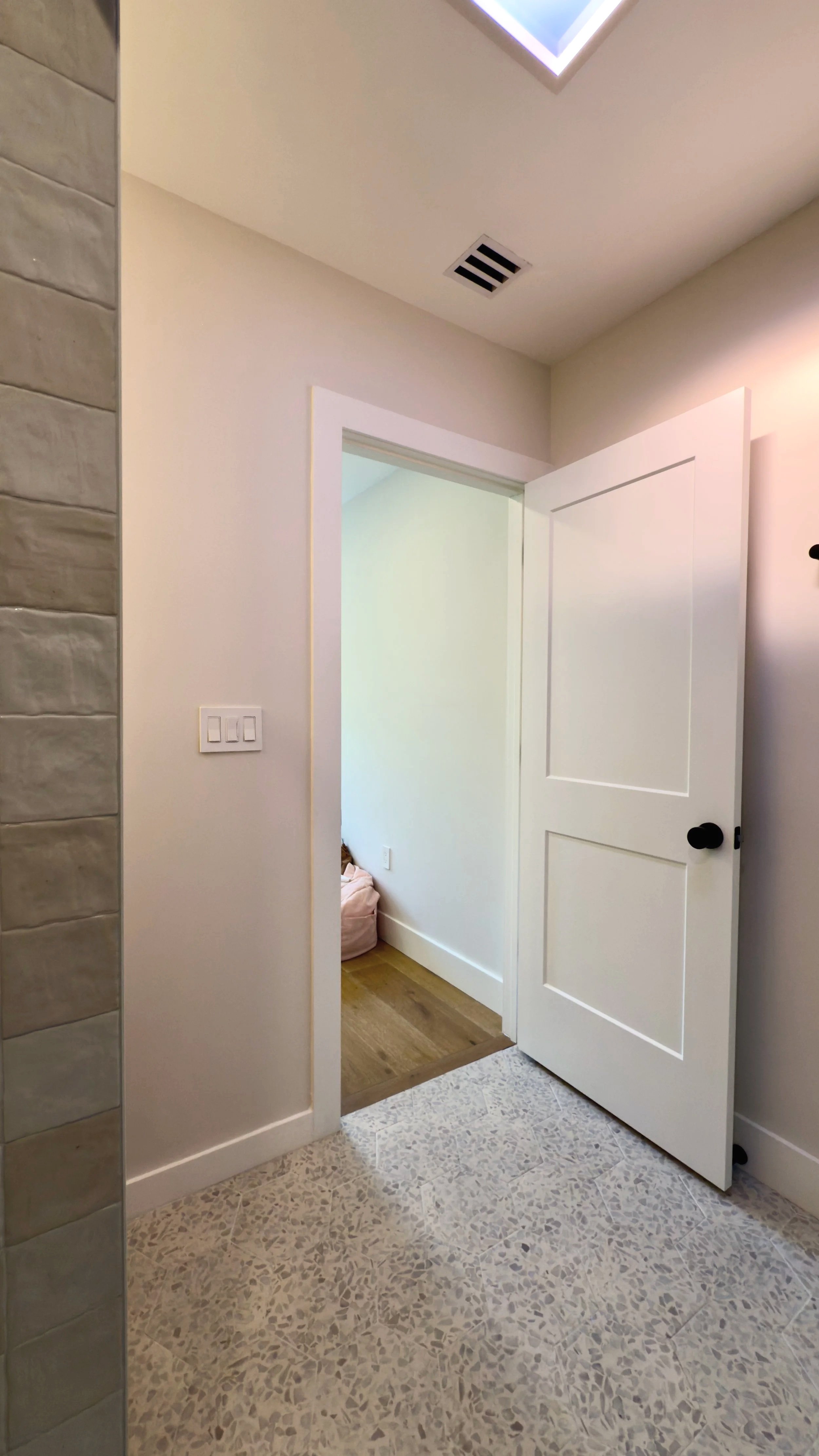 An open white door leading to a room with wood flooring, visible through a light switch area on a white wall, with a partially visible ceiling vent and a ceiling light fixture.