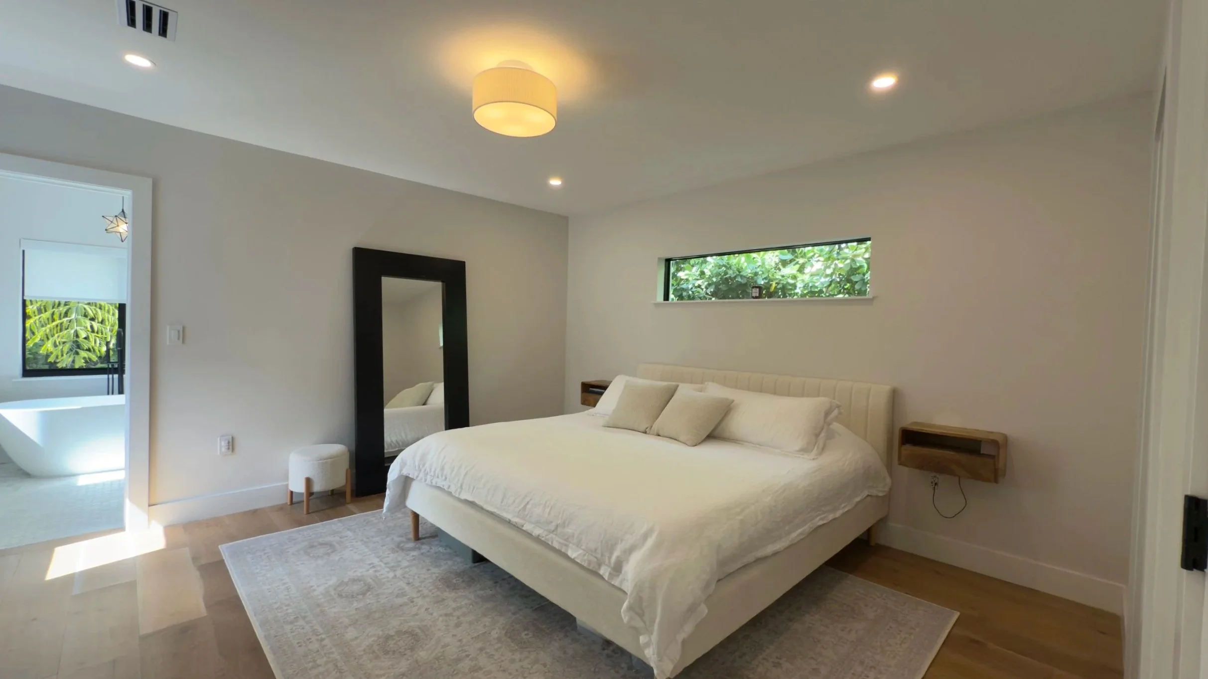 Modern bedroom with a large white bed, wooden side tables, a mirror, a window with greenery outside, and an en-suite bathroom with a bathtub visible through the open door.