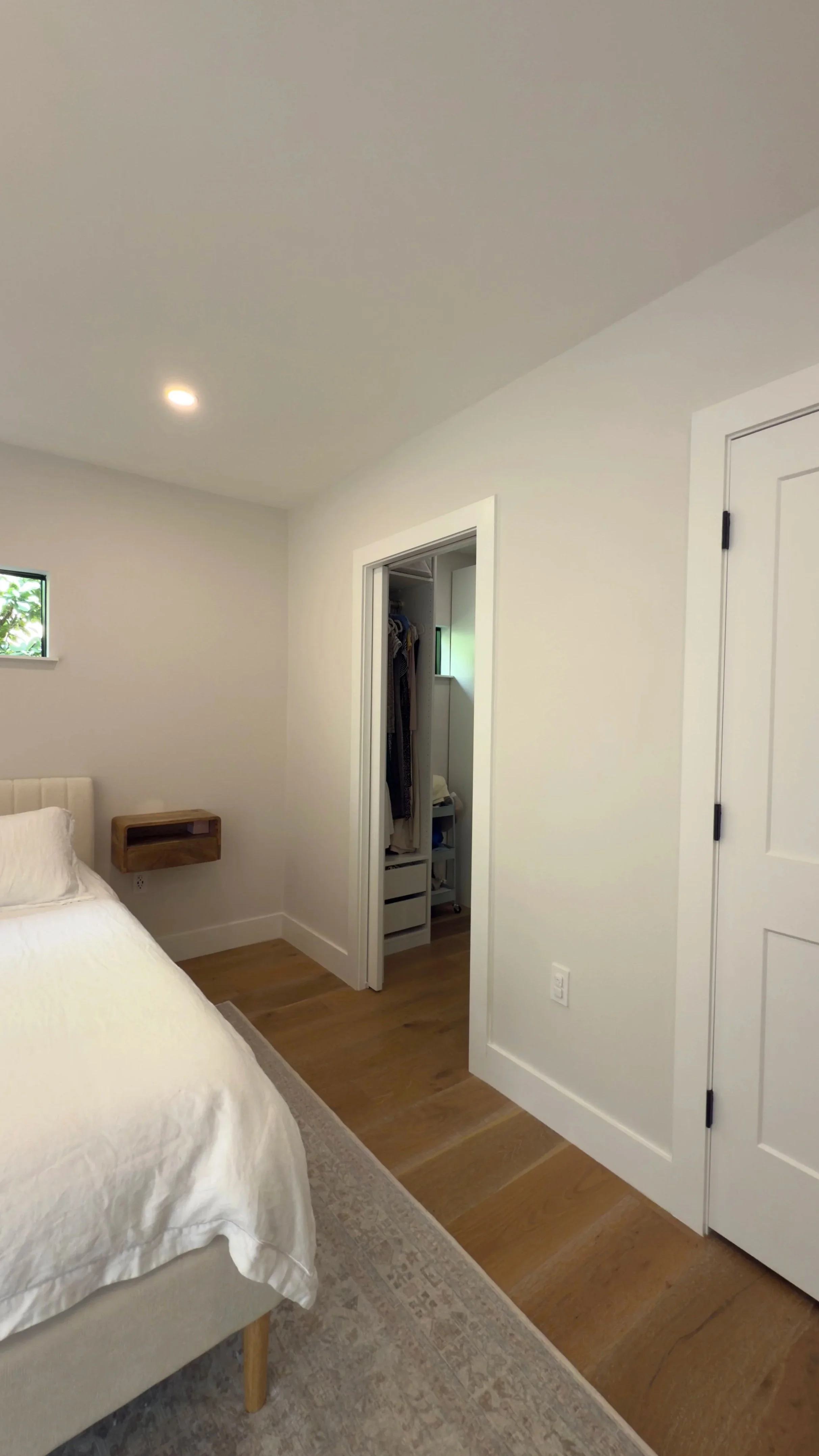 A bedroom with a white bed, wooden nightstand, small closet with hanging clothes, and a small window.