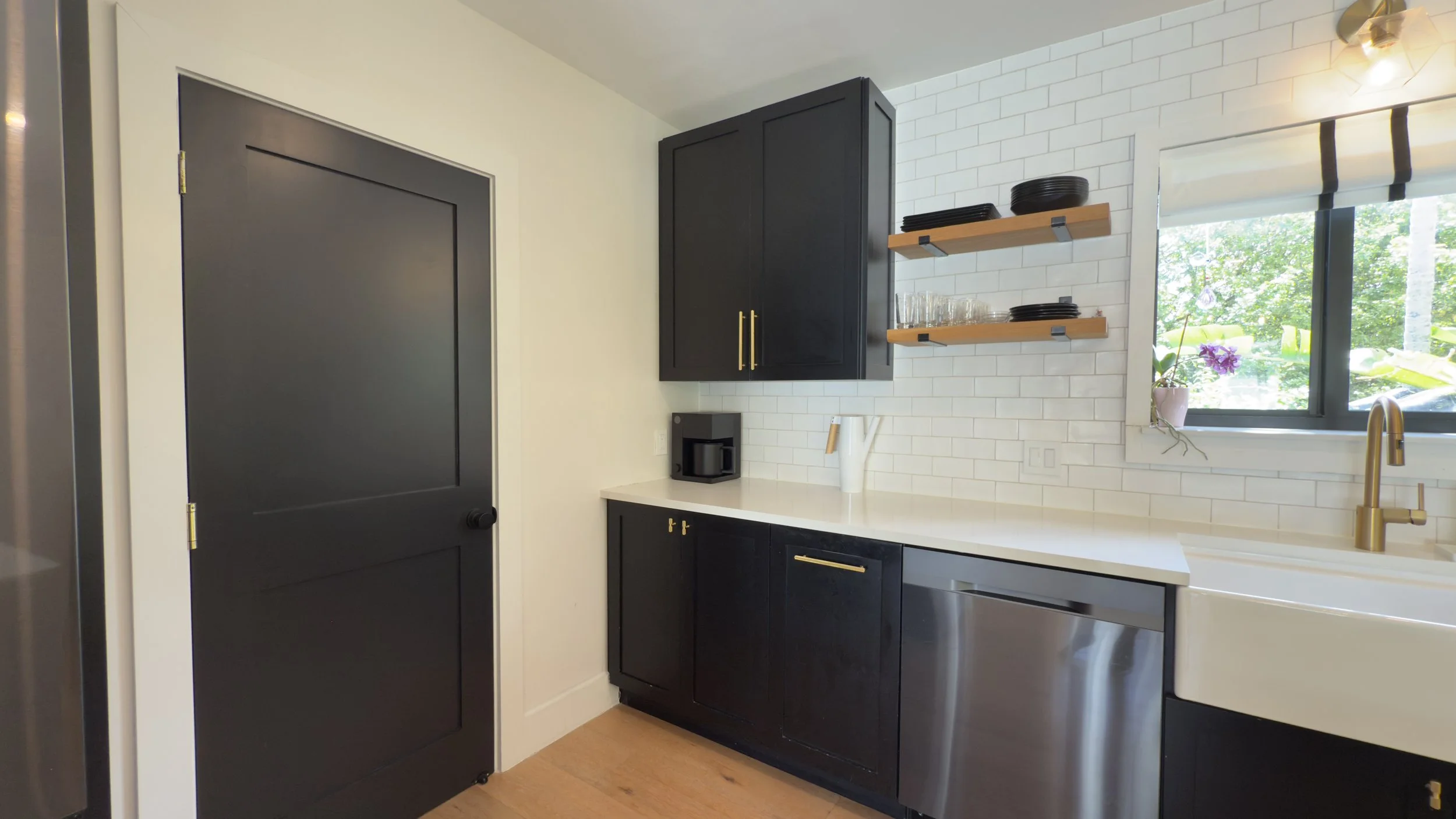 Modern kitchenette with black cabinets, white countertop, stainless steel dishwasher, and white subway tile backsplash. Open shelving holding dishes and glasses. Window with view of greenery.