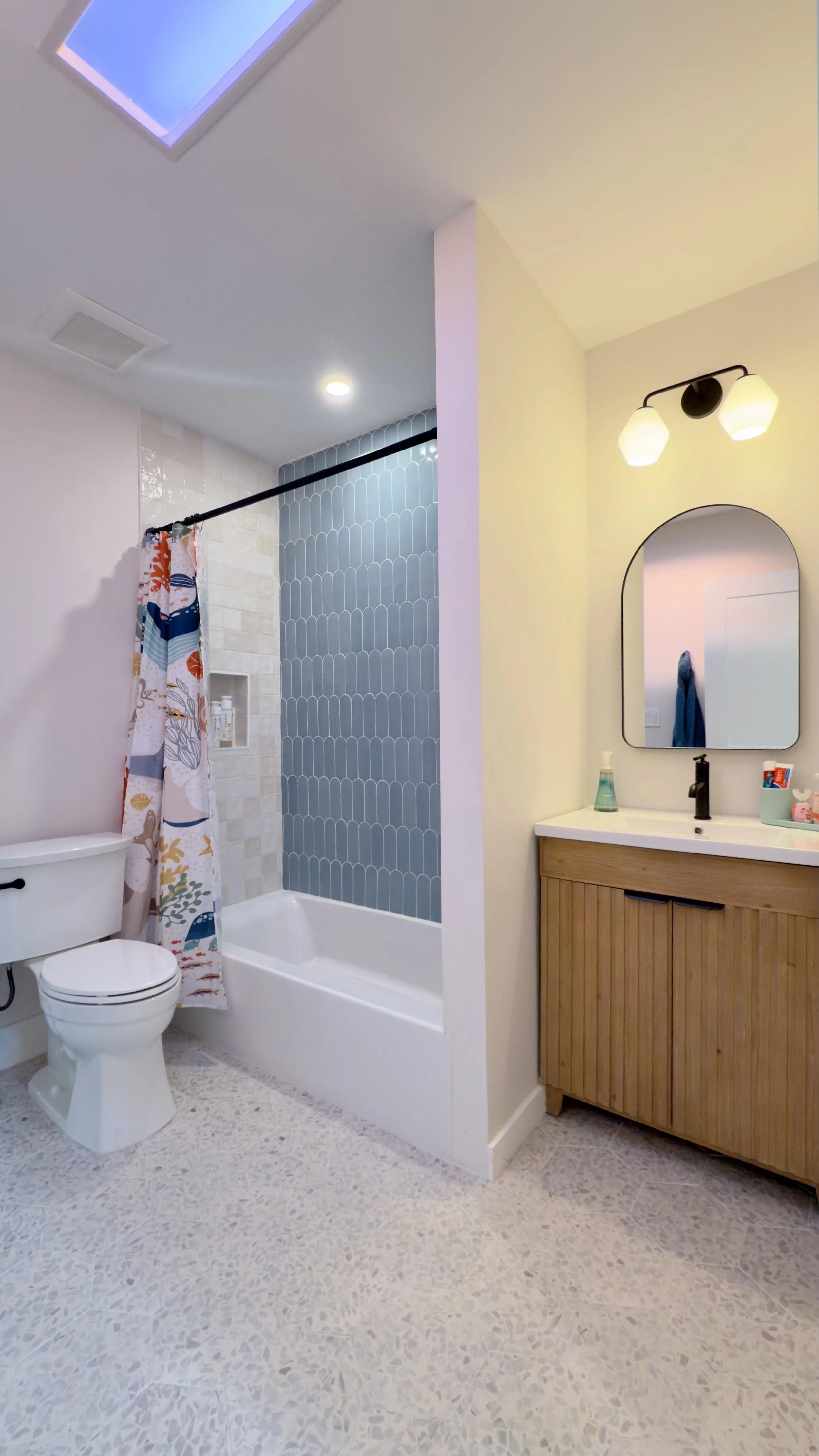 Bathroom with a toilet, bathtub with blue tile wall, wooden vanity with mirror, and colorful shower curtain.