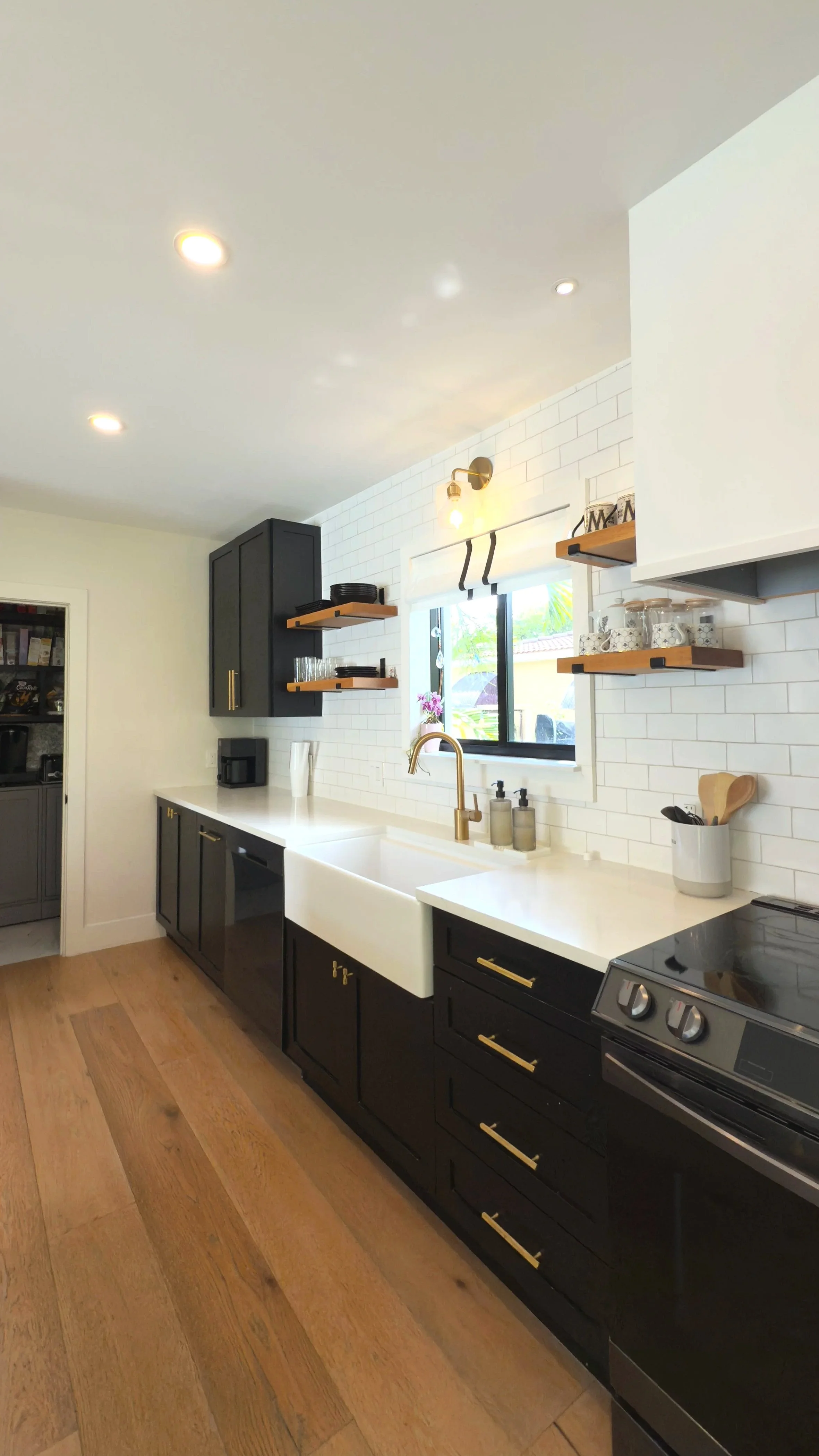 Modern kitchen with black cabinets, white farmhouse sink, white subway tile backsplash, gold accents, open wooden shelves, window, wood flooring, and black appliances.