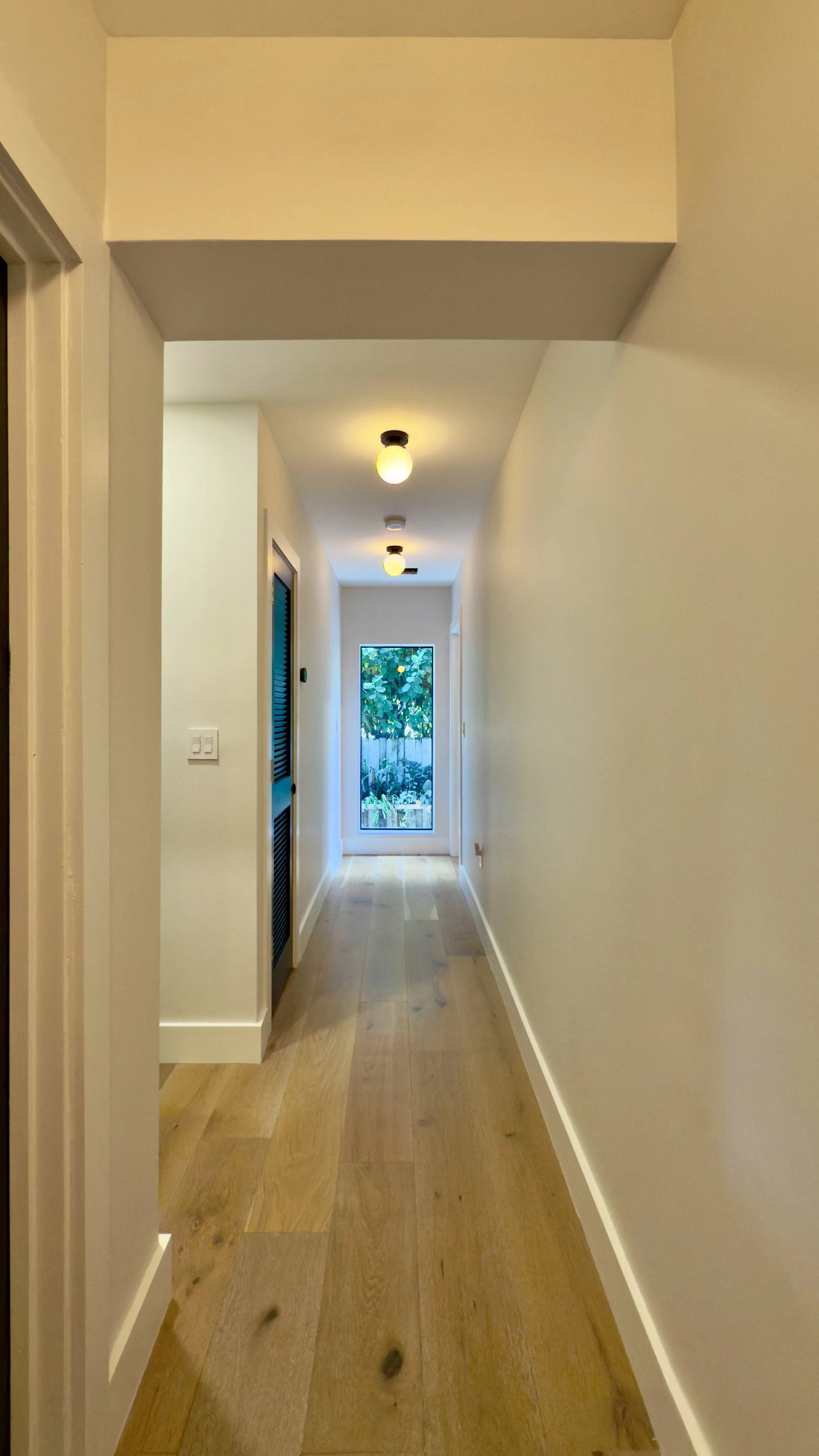 A hallway with white walls and wooden flooring, illuminated by ceiling lights, leading to a window at the end with greenery outside.