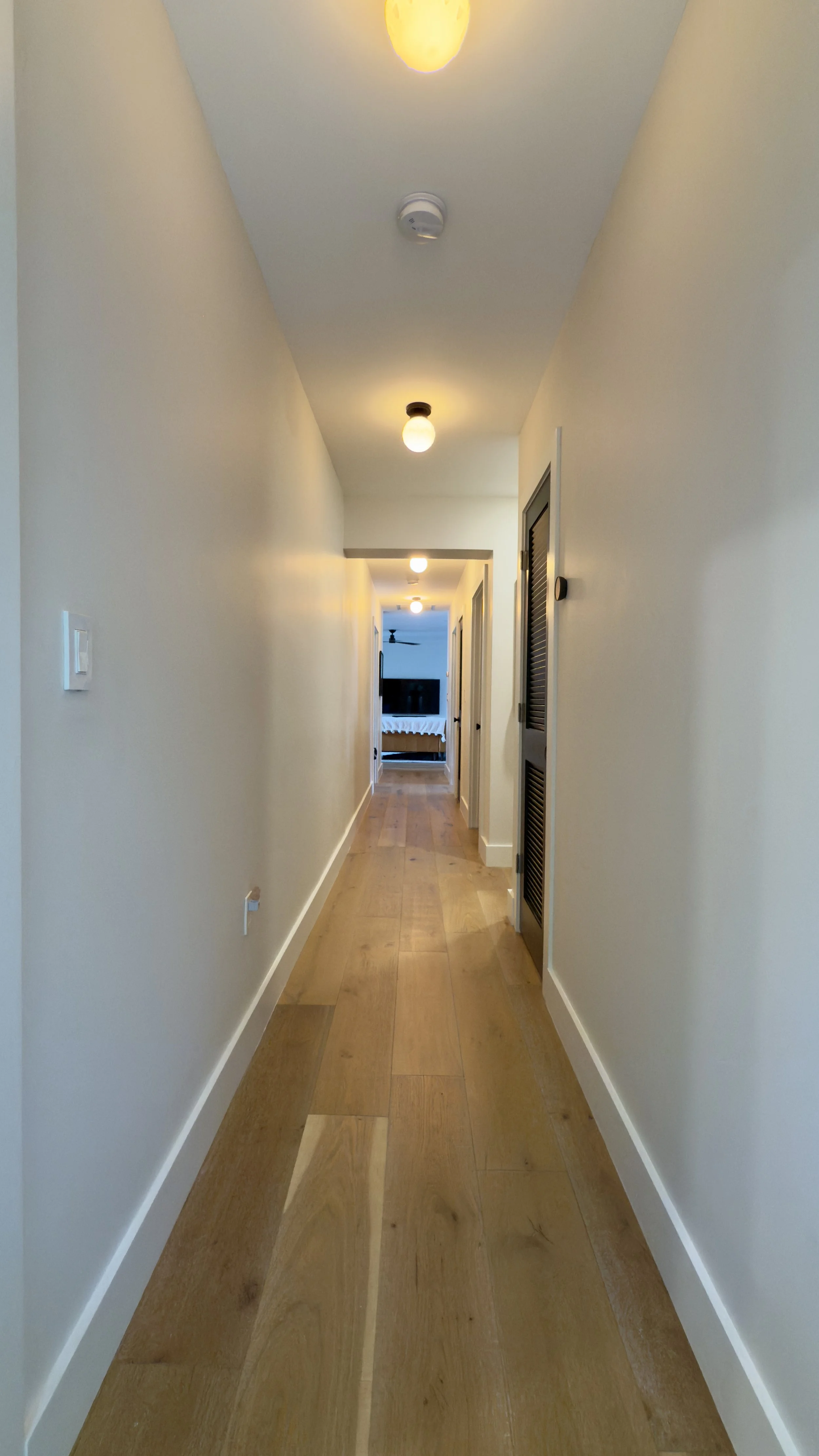 A long hallway with wooden flooring, beige walls, and ceiling lights, leading to a room with a bed and a television.