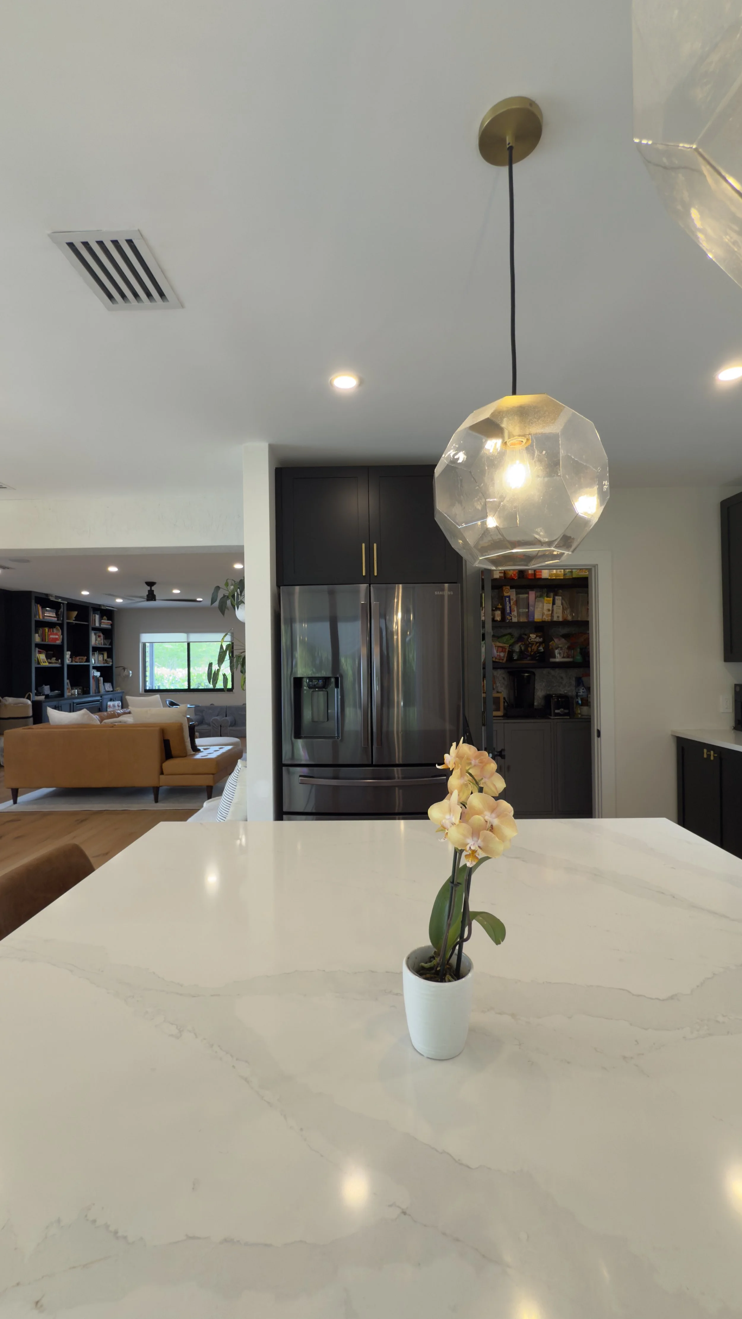 A kitchen with a white marble countertop, a small potted orchid plant, a black pendant light, a stainless steel refrigerator, and a view into a living room with bookshelves and a sofa.