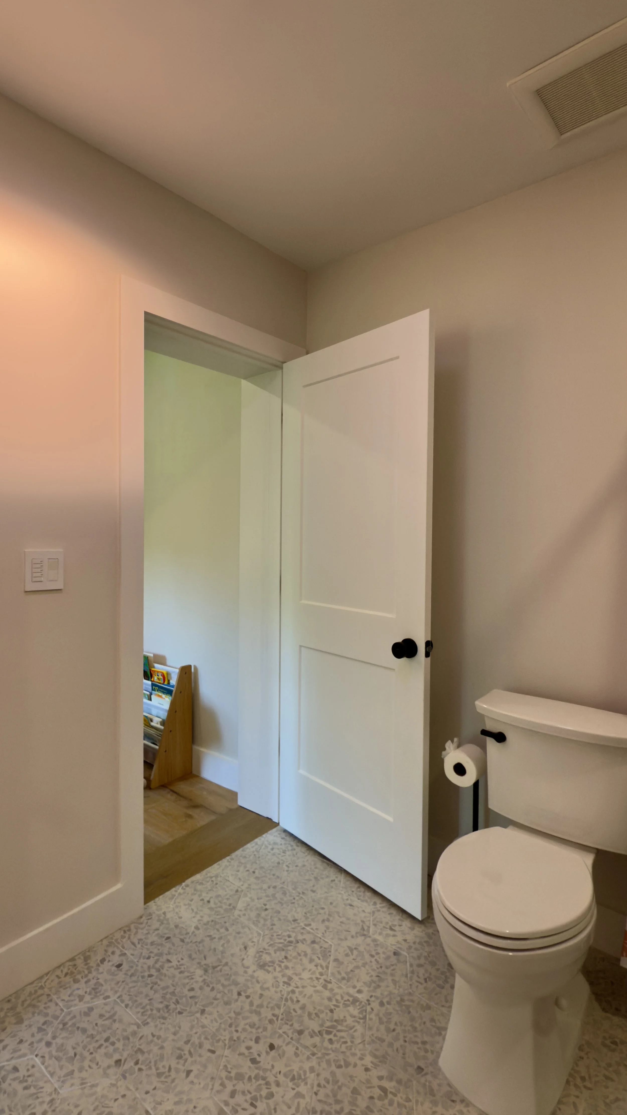A bathroom with beige tiled flooring, a white toilet, and a partially open white door leading to a children's toy area.