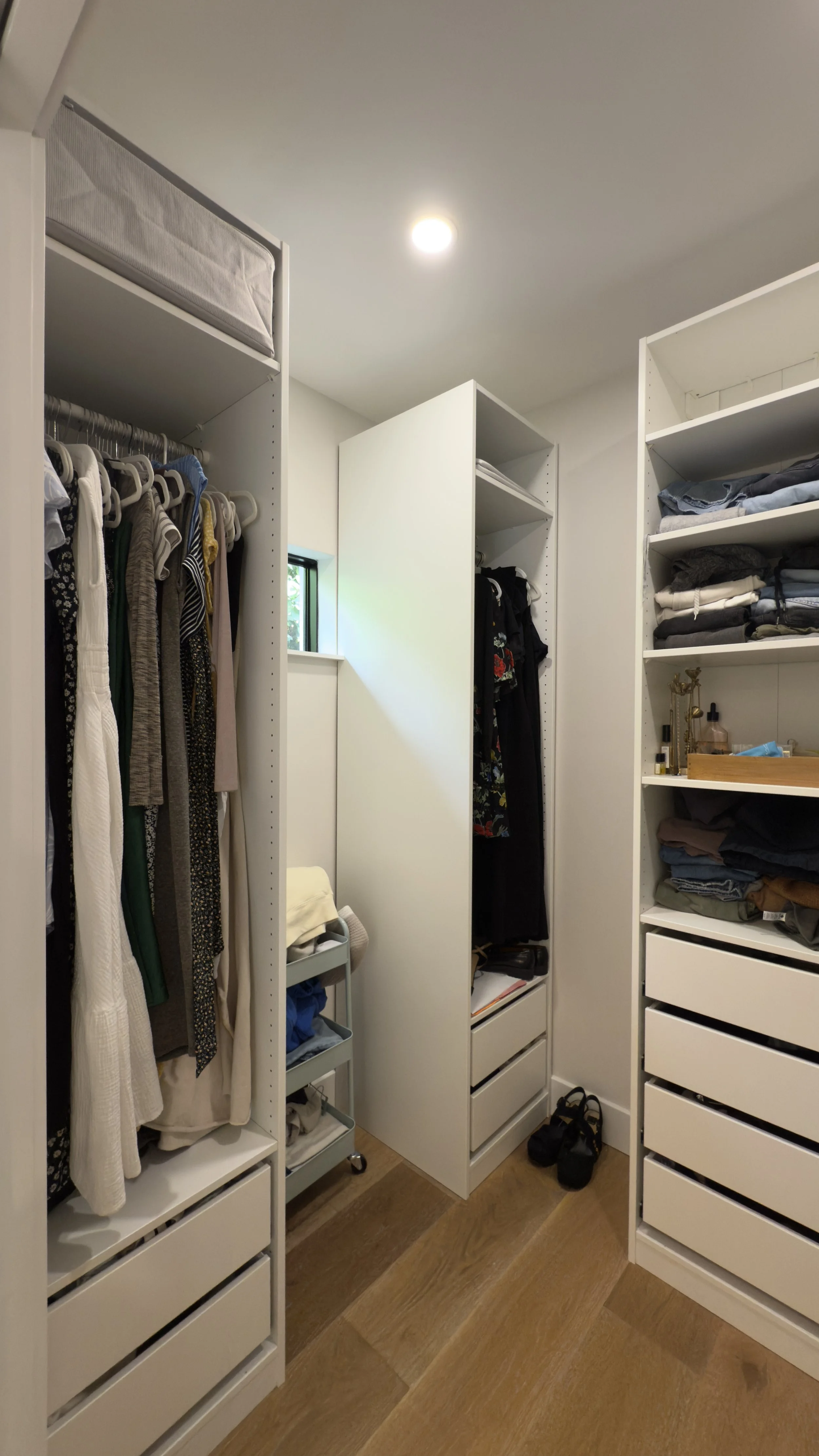 A walk-in closet with white shelving units, hanging clothes, folded laundry, a small window, and a pair of black shoes on wooden flooring.