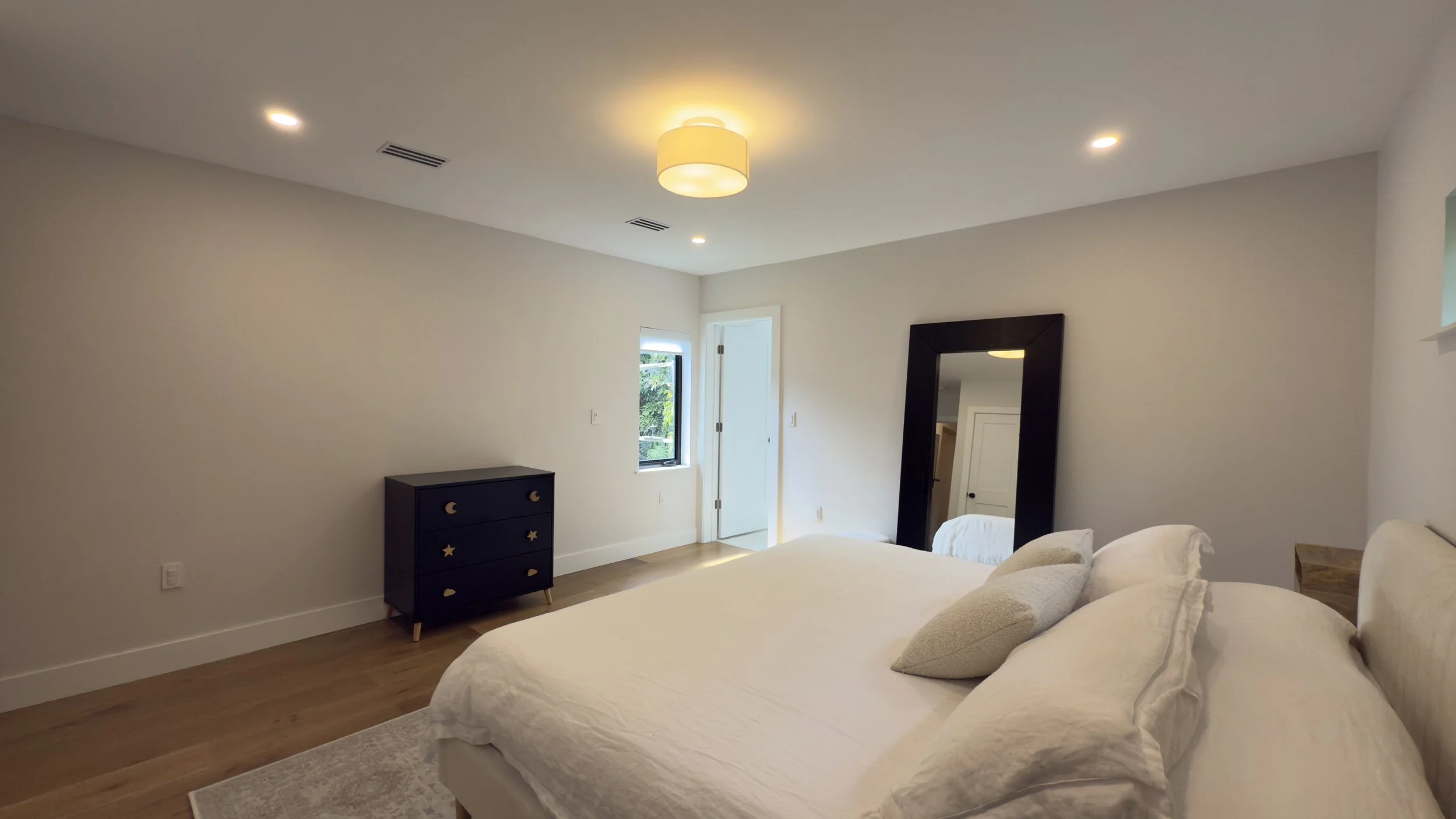 A bedroom with a bed, pillows, a full-length mirror, a window, and a dresser with moon and star decorations in a minimalist decor style.