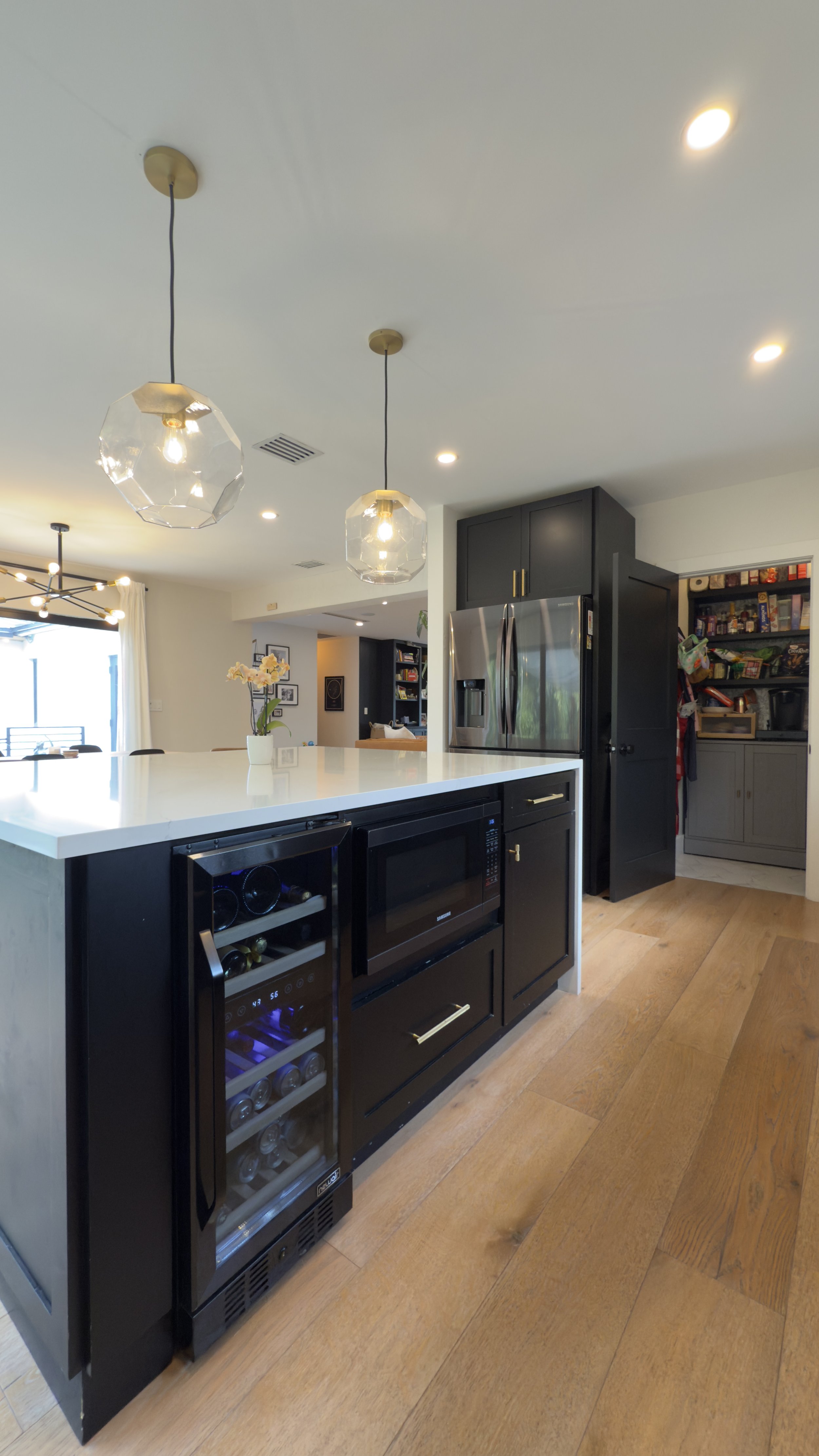 Modern kitchen with black cabinetry, stainless steel refrigerator, built-in microwave, wine fridge, and pendant lighting over white island.