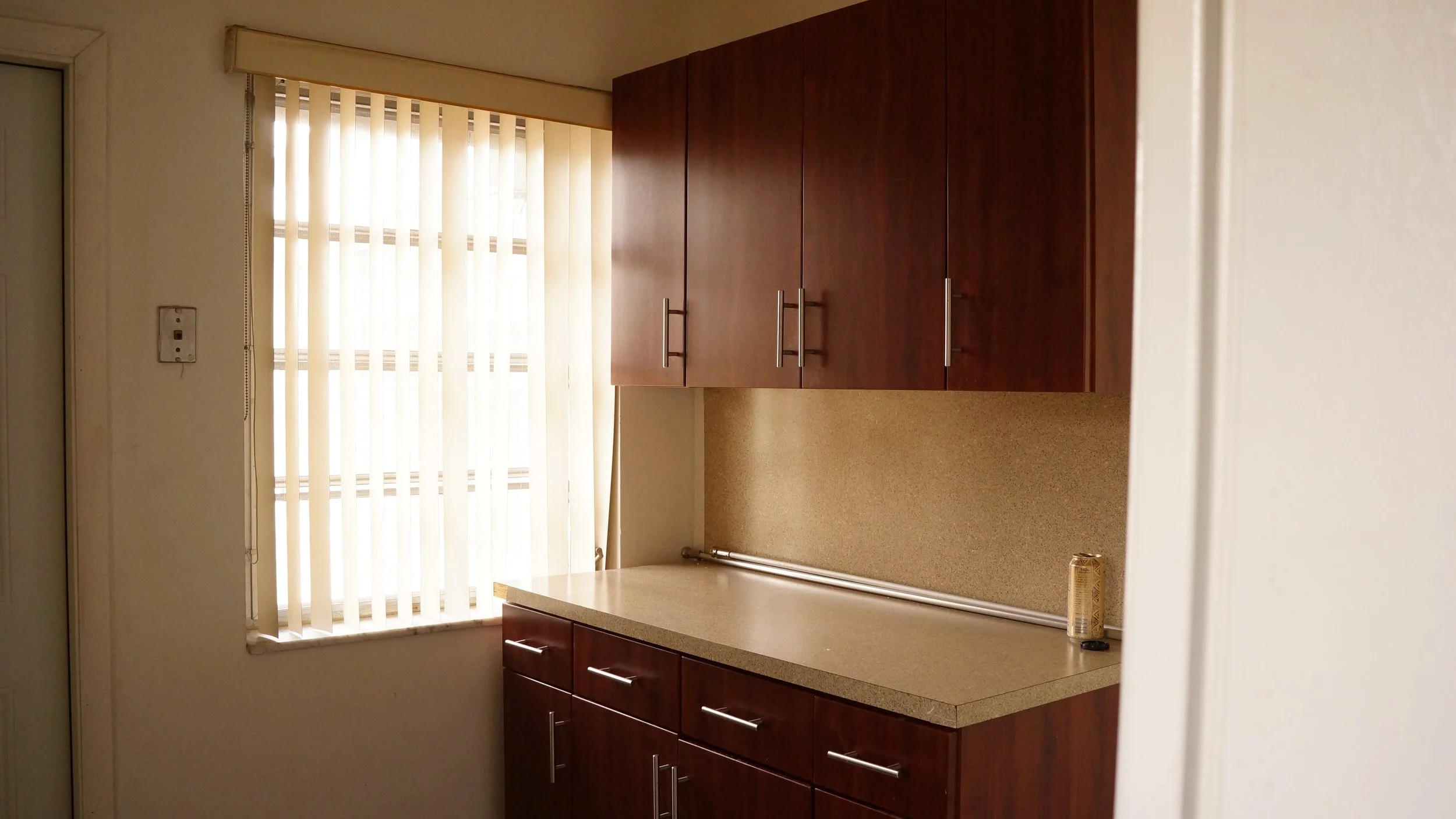 Kitchen with wooden cabinets, beige countertop, a window with vertical blinds, and a single can on the counter.