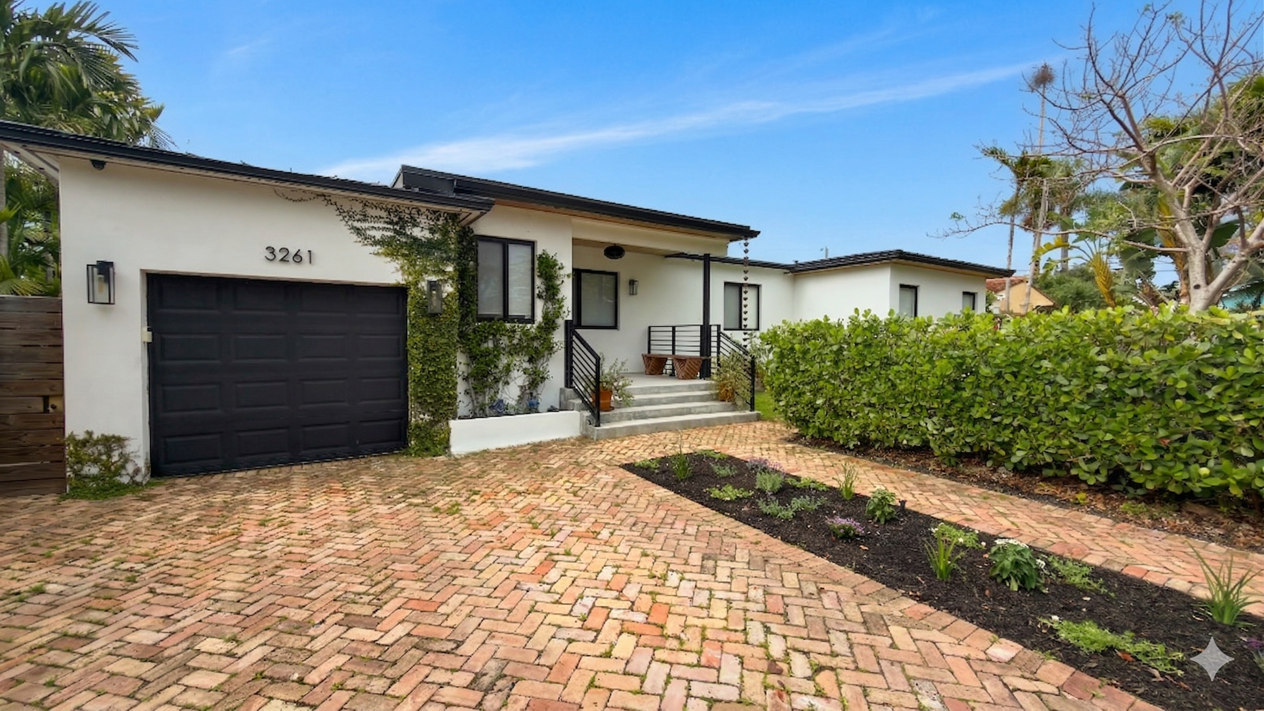 Modern single-story house with white exterior, black garage door, small front porch with stairs, and landscaped front yard with brick driveway and garden bed.