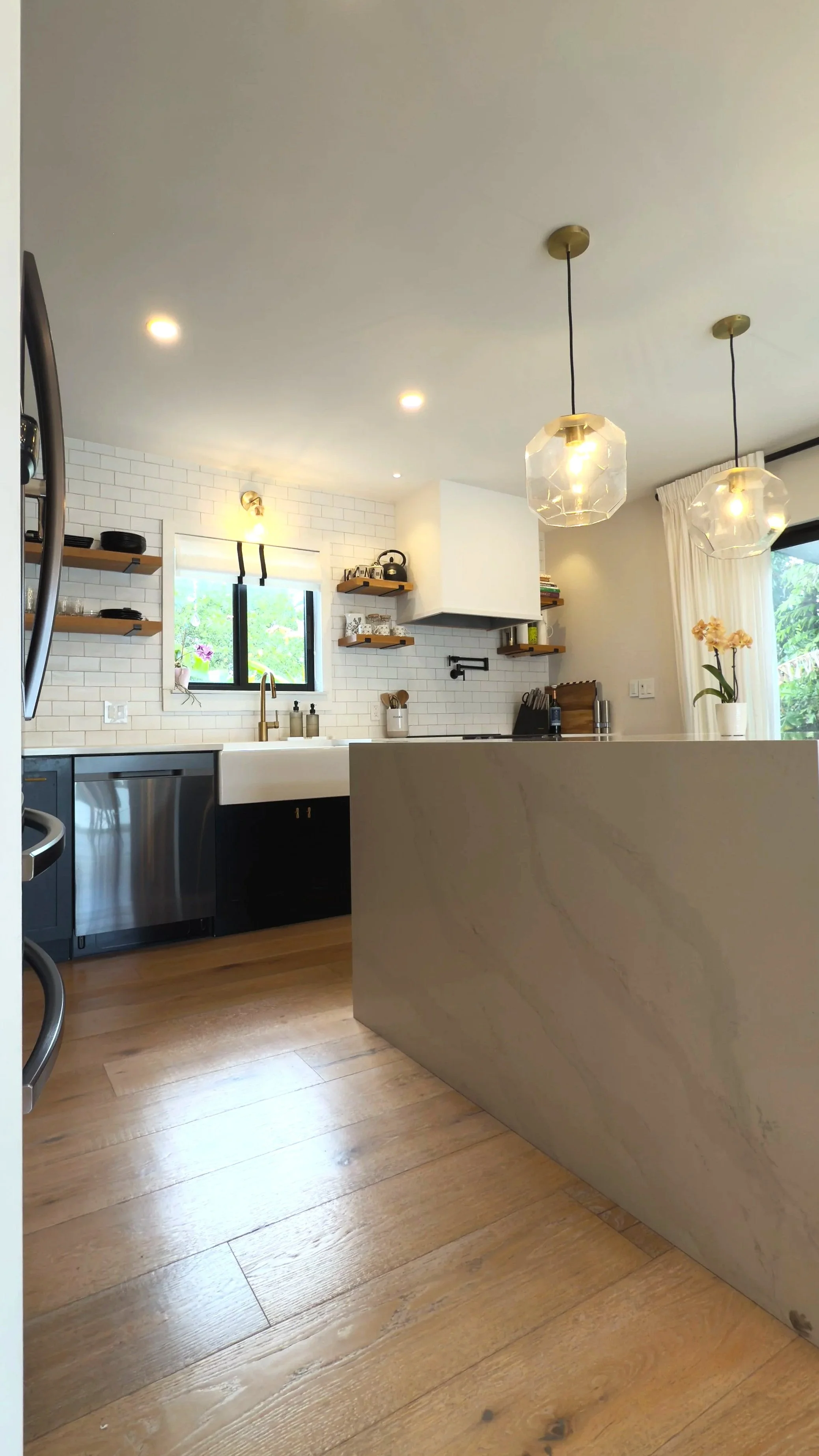 Modern kitchen with white subway tile backsplash, wooden open shelves, black and stainless steel appliances, a window above the sink, and pendant lights hanging over a marble kitchen island.