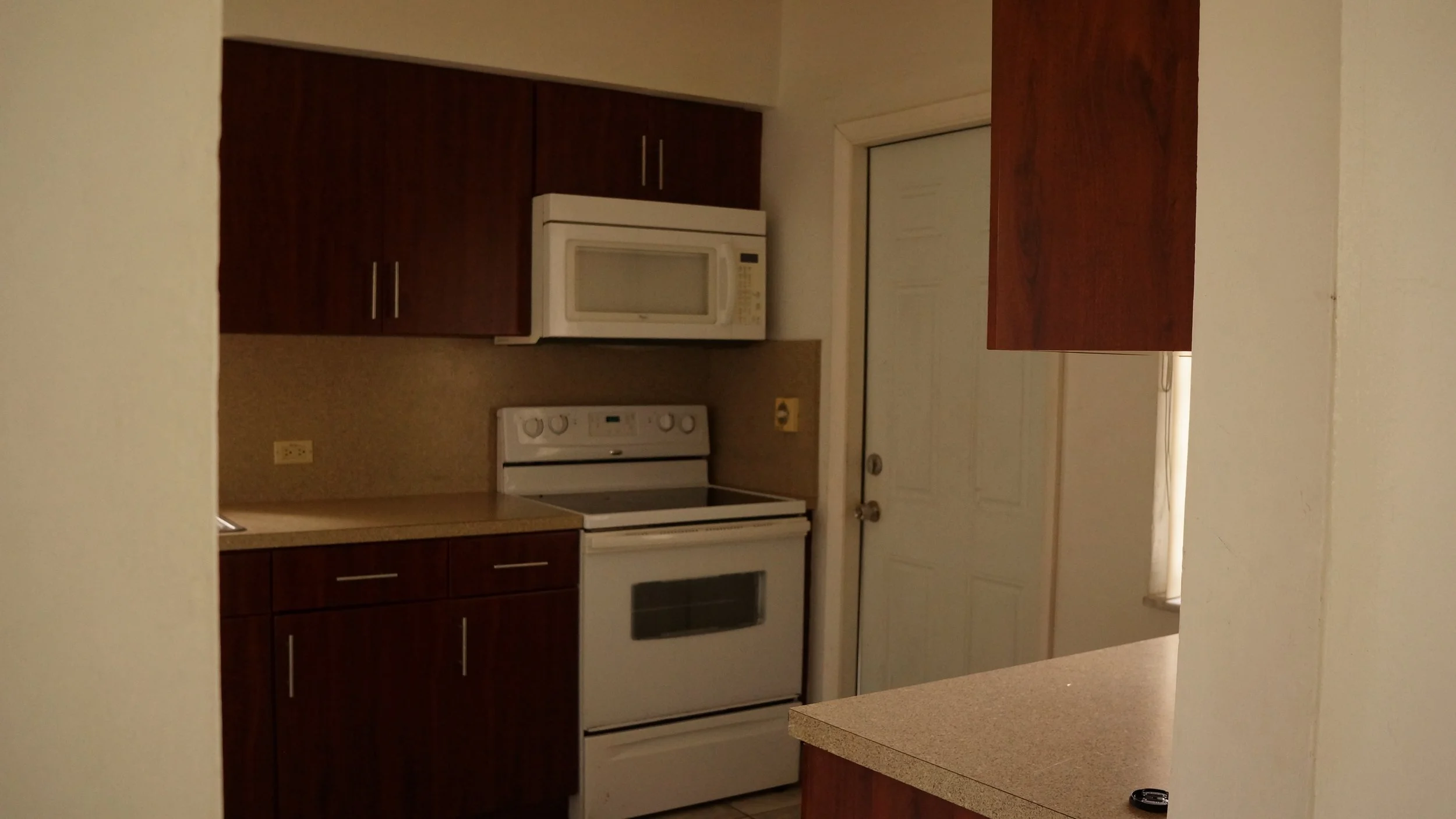 View of a small kitchen with dark wood cabinets, a white microwave, a white stove, and beige countertops, seen through a partial wall opening.
