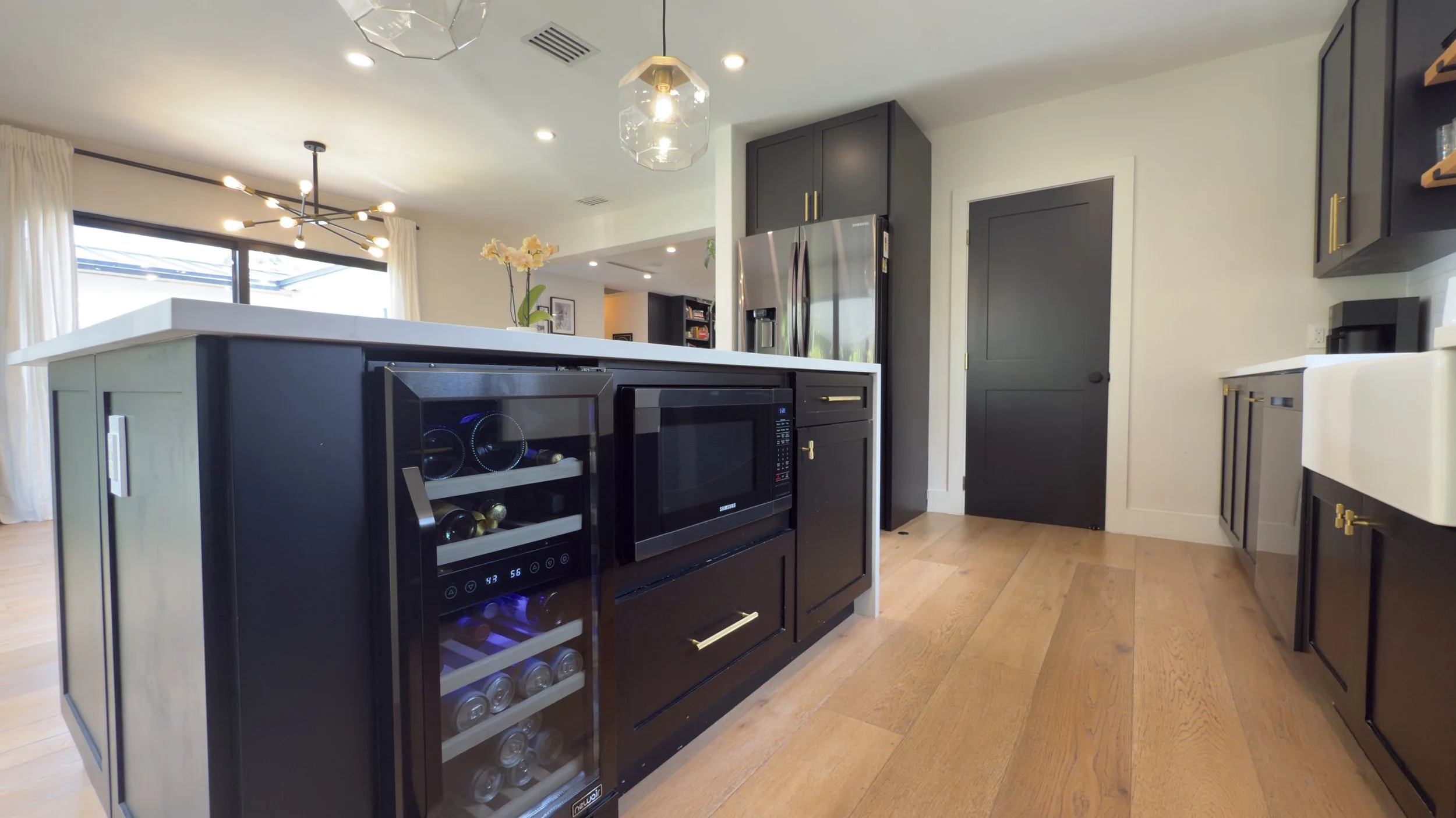 Modern kitchen with black cabinets, white countertops, stainless steel refrigerator, black microwave, wine fridge, hardwood floors, and a sliding door.