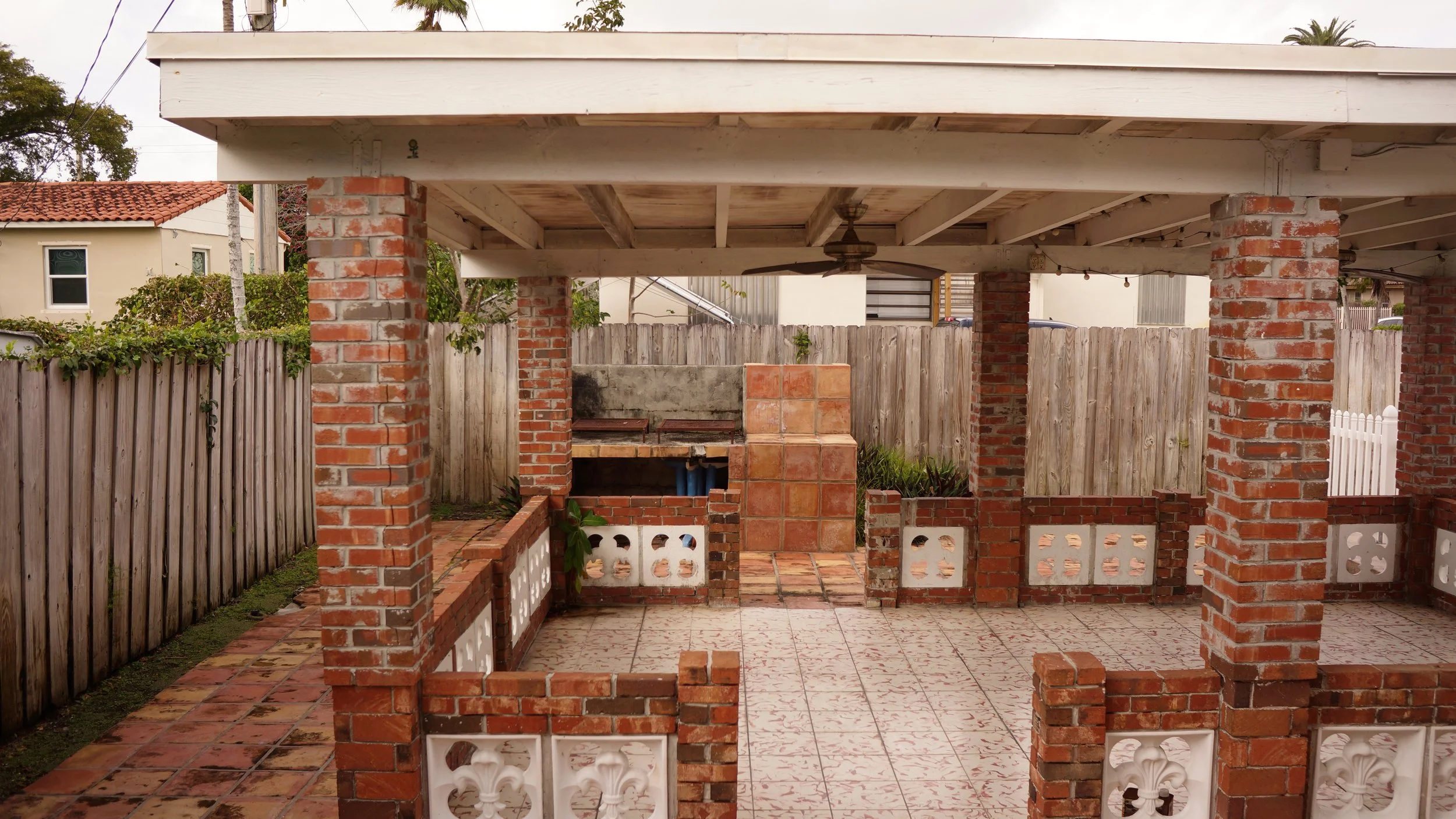 An outdoor patio with brick columns and a tiled floor, partially enclosed with a wooden fence, featuring a built-in brick and tile barbecue area under a roof with a ceiling fan.