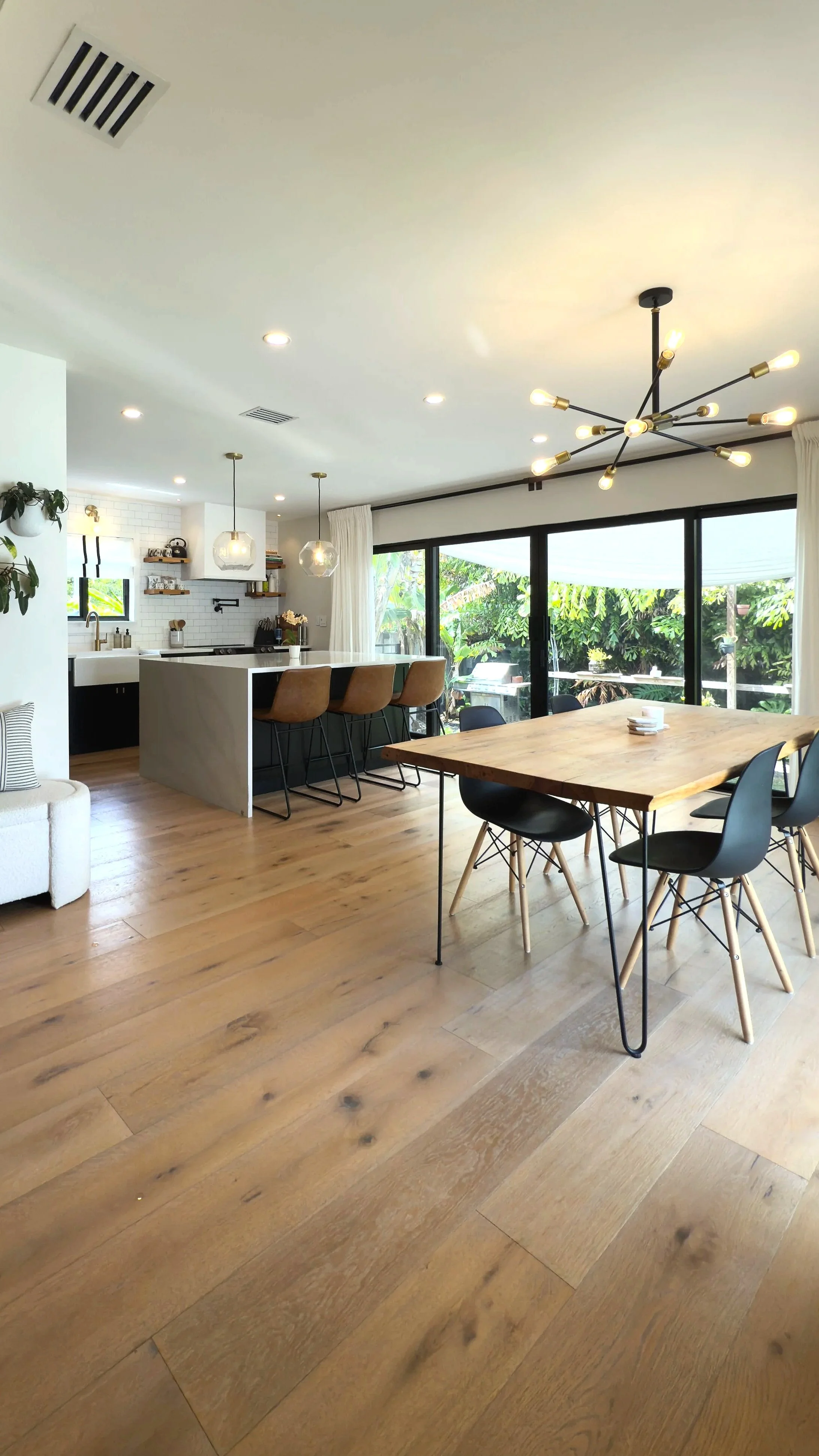 Open-concept kitchen and dining area with wooden floors, a wooden dining table with black chairs, a kitchen island with tan bar stools, pendant lights, large windows, and a modern black chandelier.