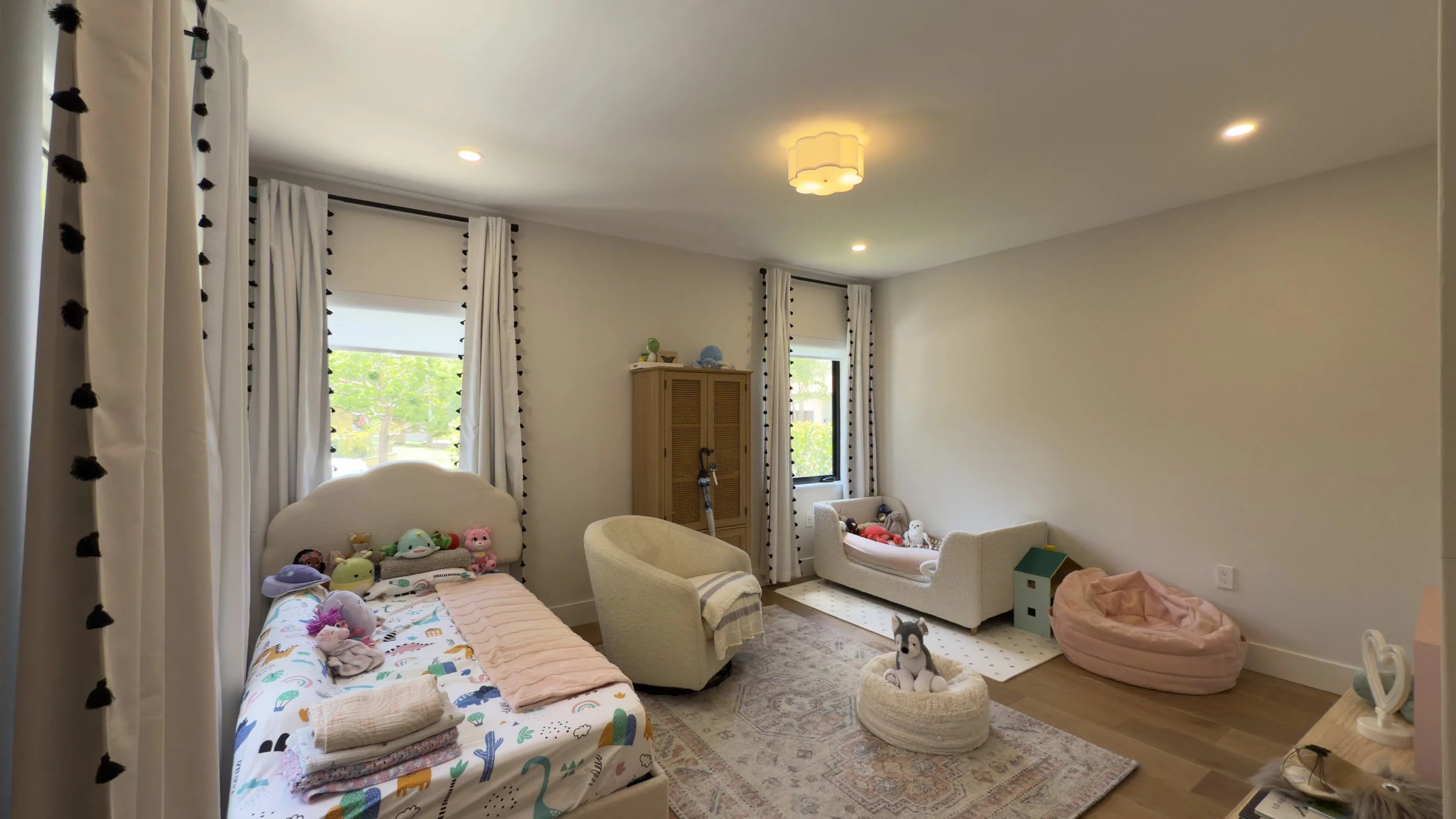 A children's bedroom with two beds, one with children's toys and stuffed animals, and a cozy nook with a bean bag and dolls. There are two windows with white curtains and black pom-pom trim, a beige armchair, and a wooden cabinet. The room has a patt