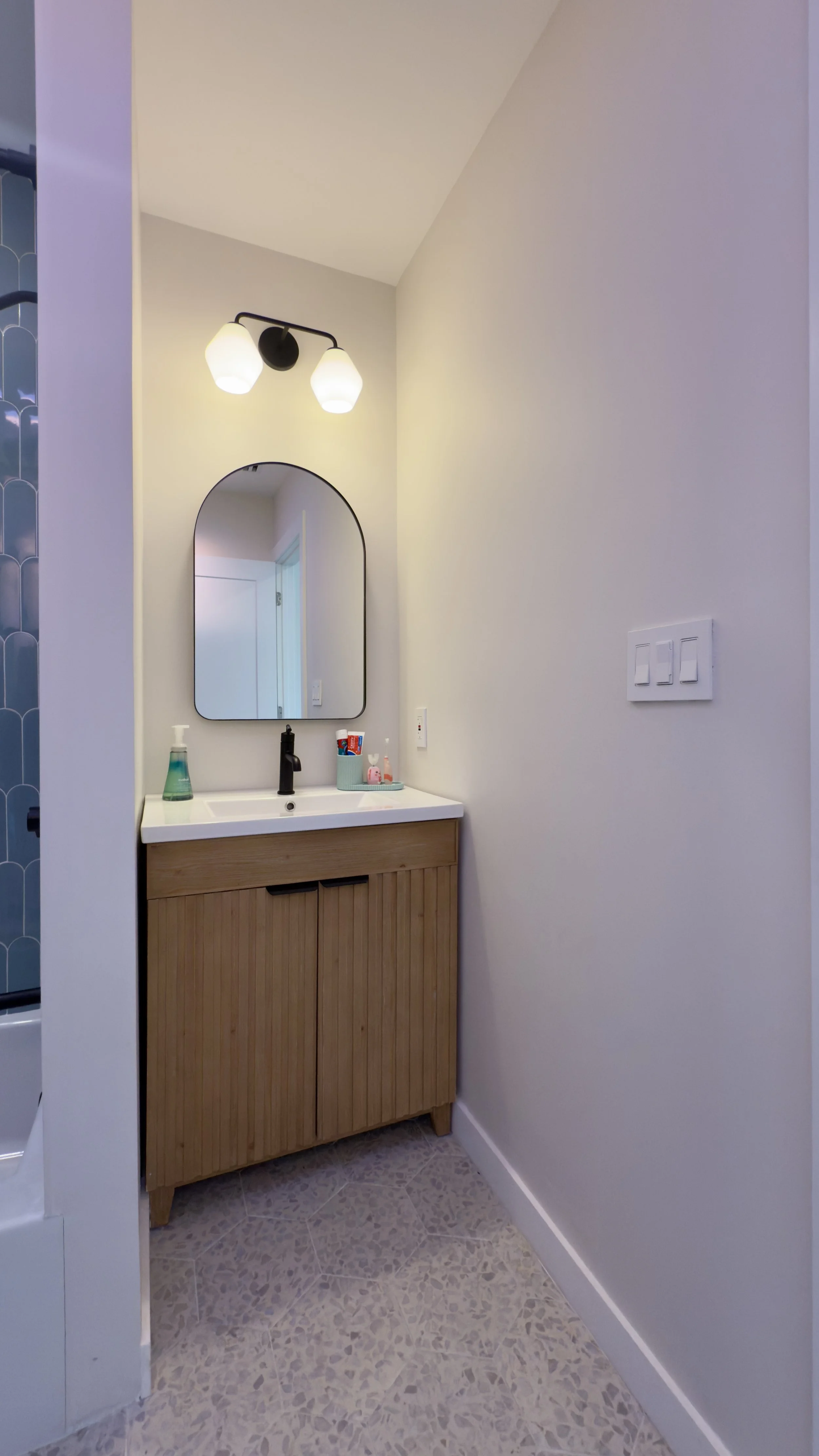 A small bathroom vanity with a wooden cabinet, a white sink, a black faucet, a mirror with rounded top, wall-mounted light fixture with two white globes, and a soap dispenser on the left side of the sink.