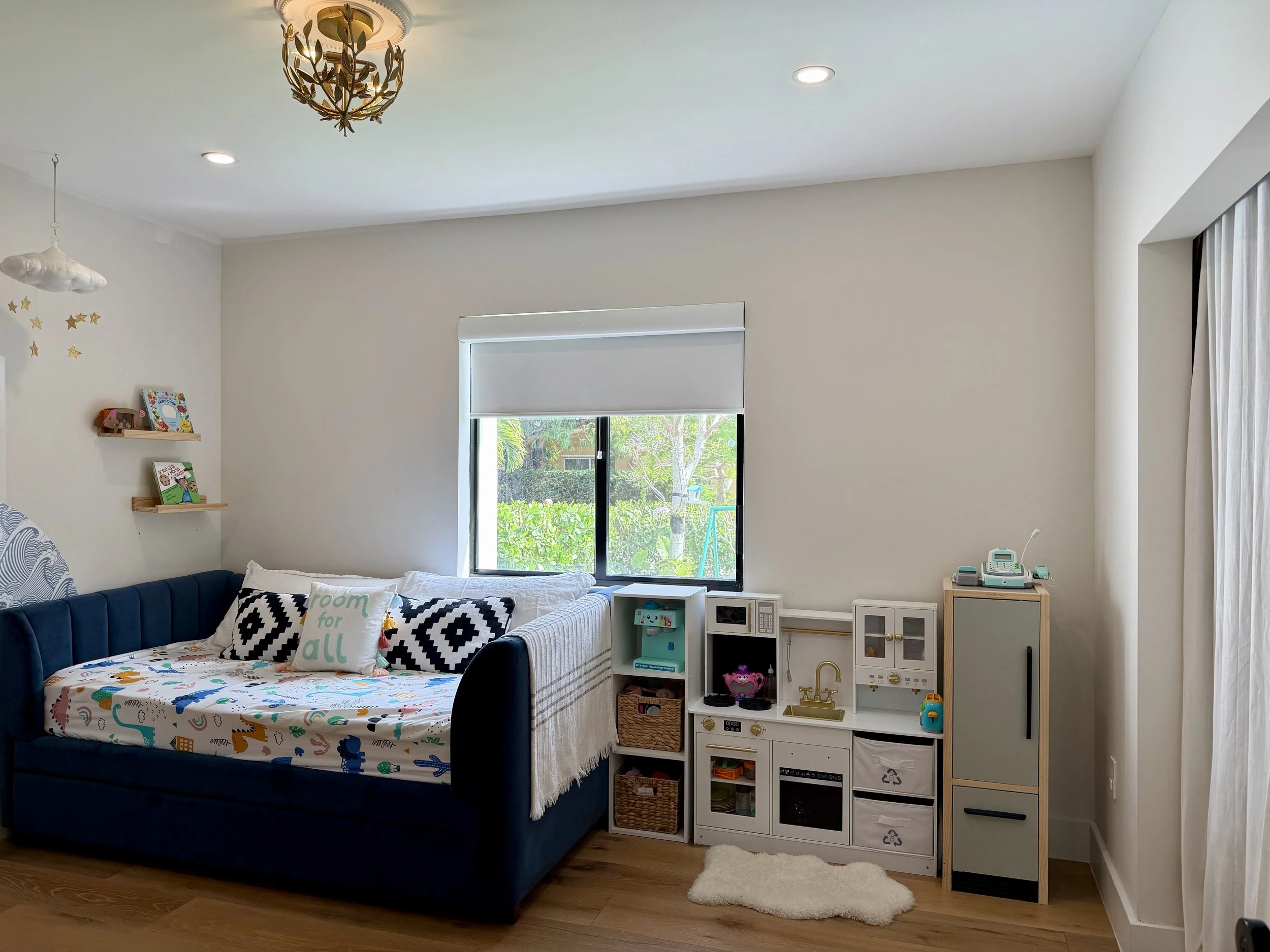 A children's bedroom with a navy blue bed featuring a playful dinosaur print bedsheet and black and white geometric pillows, white shelves with books and toys on the wall, a window with a view of greenery, and a white toy kitchen set with a small rug