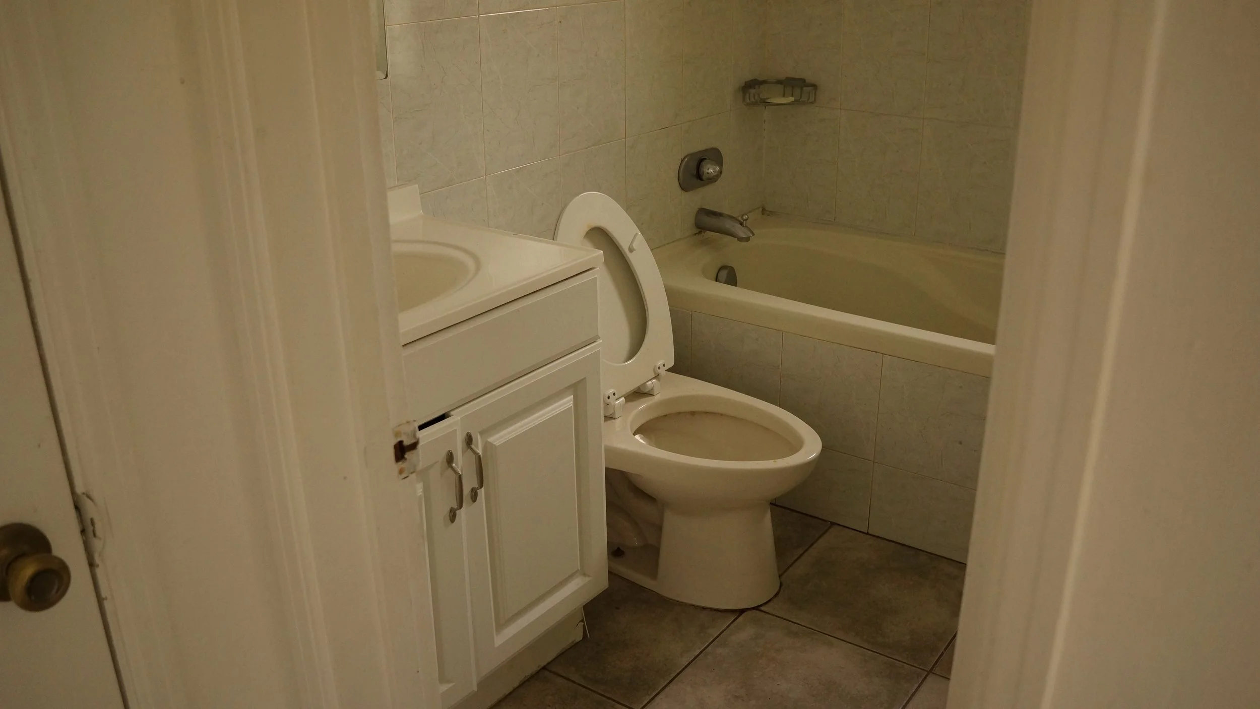 A bathroom with a white toilet, a white sink cabinet, and a bathtub with a wall-mounted soap holder, all in a small tiled room.