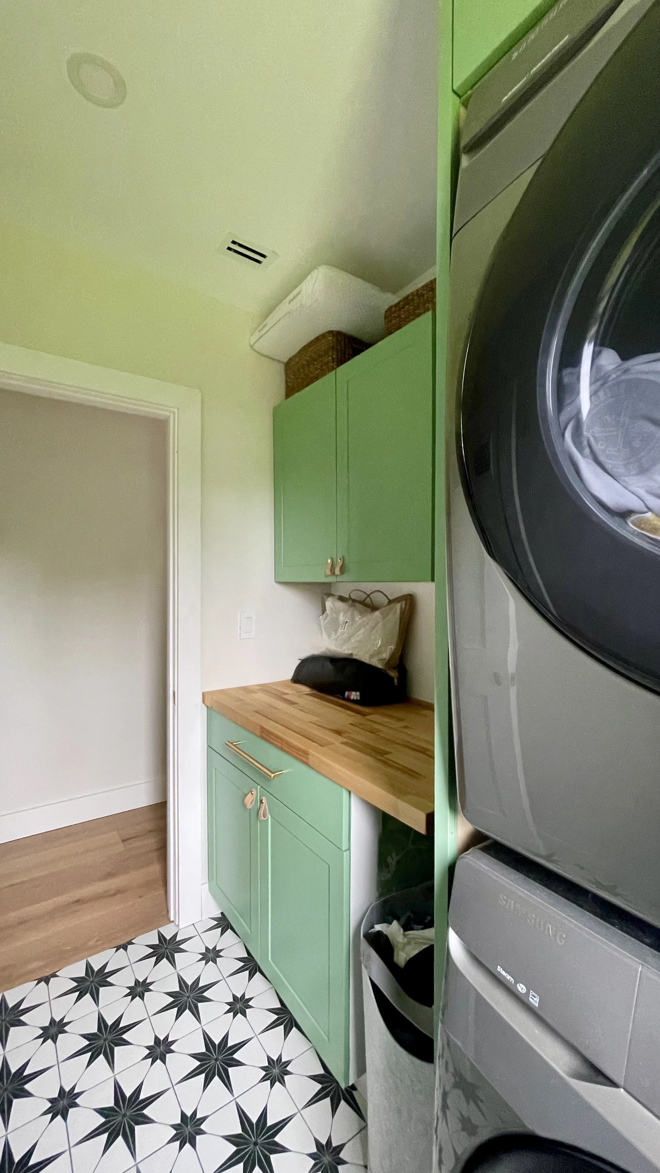 A laundry room with a black washing machine on the right, green cabinets above and below a wooden countertop, patterned black and white tile floor, and a wicker basket and a white item on top of the cabinets.