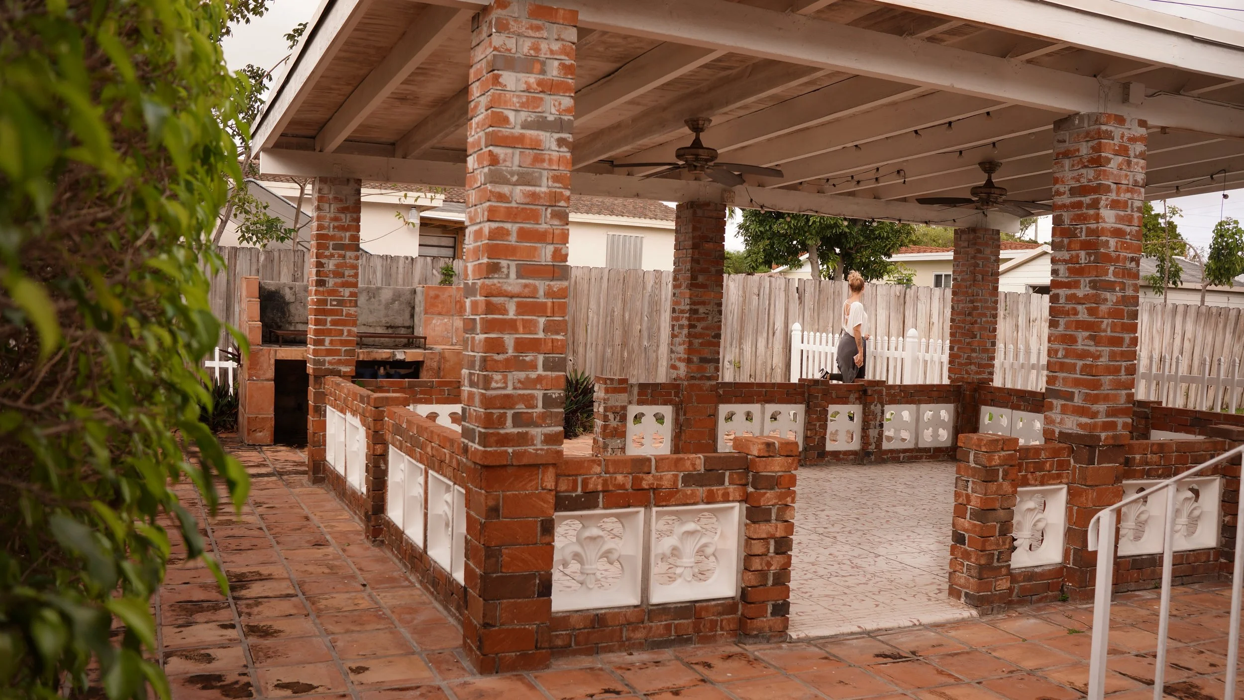 A covered brick patio with a white decorative banister, brick columns, and ceiling fans, with a woman standing at the fence and a barbecue grill in the corner.