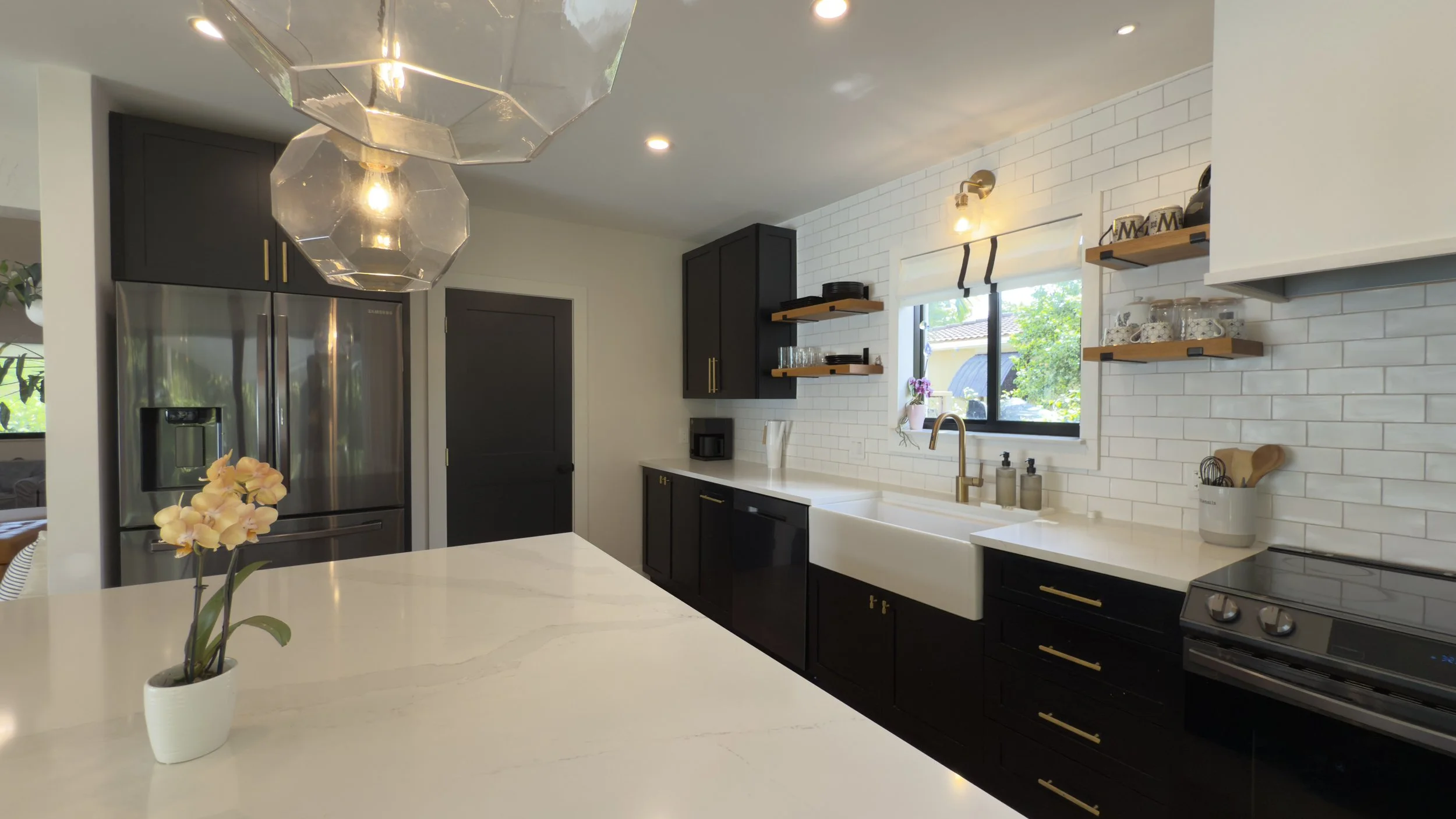 Modern kitchen with white countertops, black cabinets with gold handles, white subway tile backsplash, farmhouse sink, stainless steel refrigerator, and floating wooden shelves with dishes and glassware. Decorative purple flowers on the window sill.