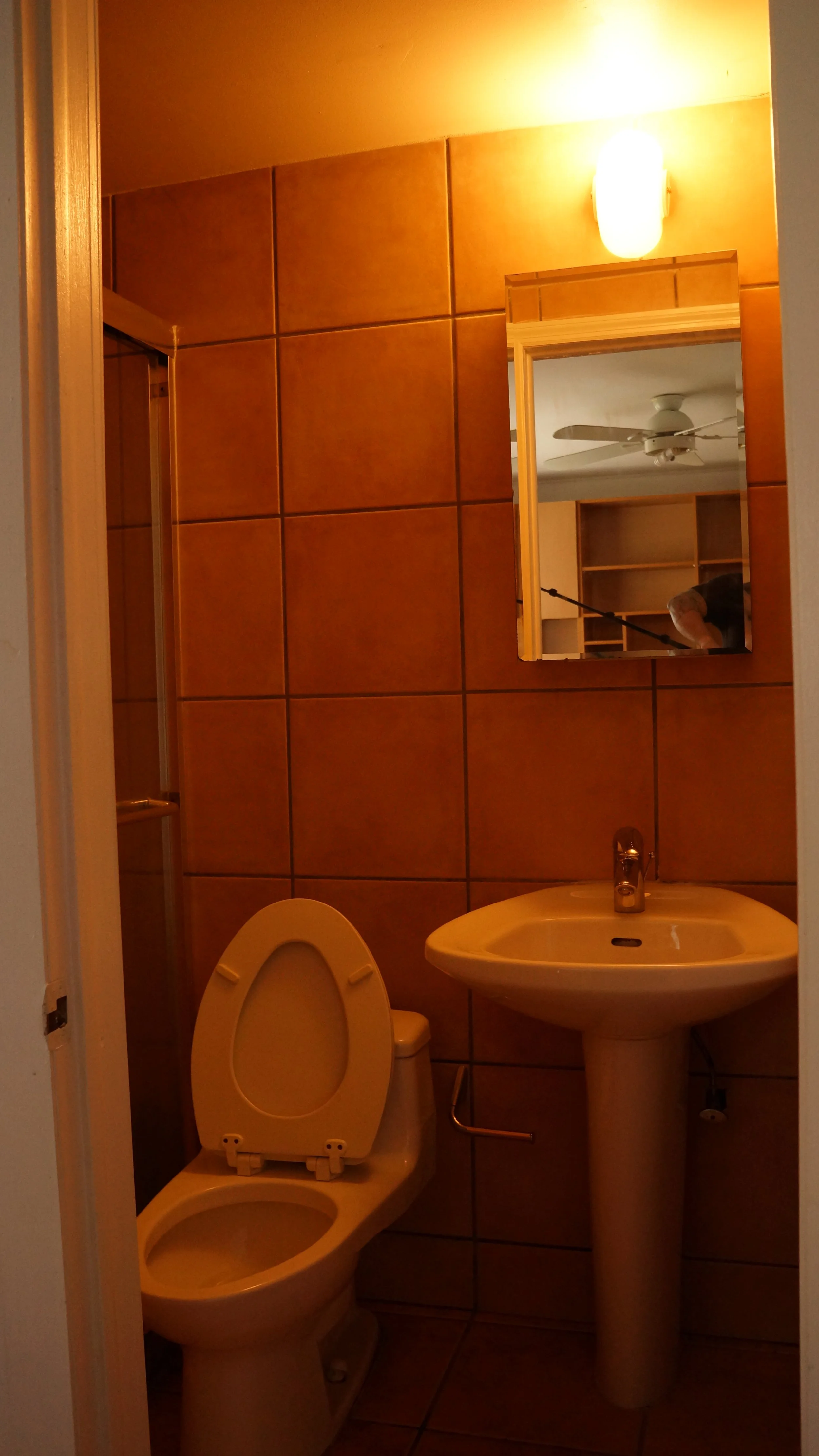 Small bathroom with beige tiles, a white toilet with the lid open, a white sink with a silver faucet, a rectangular mirror above the sink, and a wall-mounted light fixture above the mirror. The bathroom door is partially open, showing a room with a c
