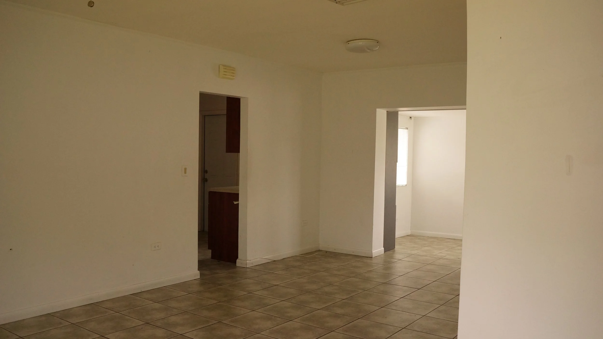 Empty room with beige tiled floor, white walls, and doorways leading to other rooms, with natural light coming in.
