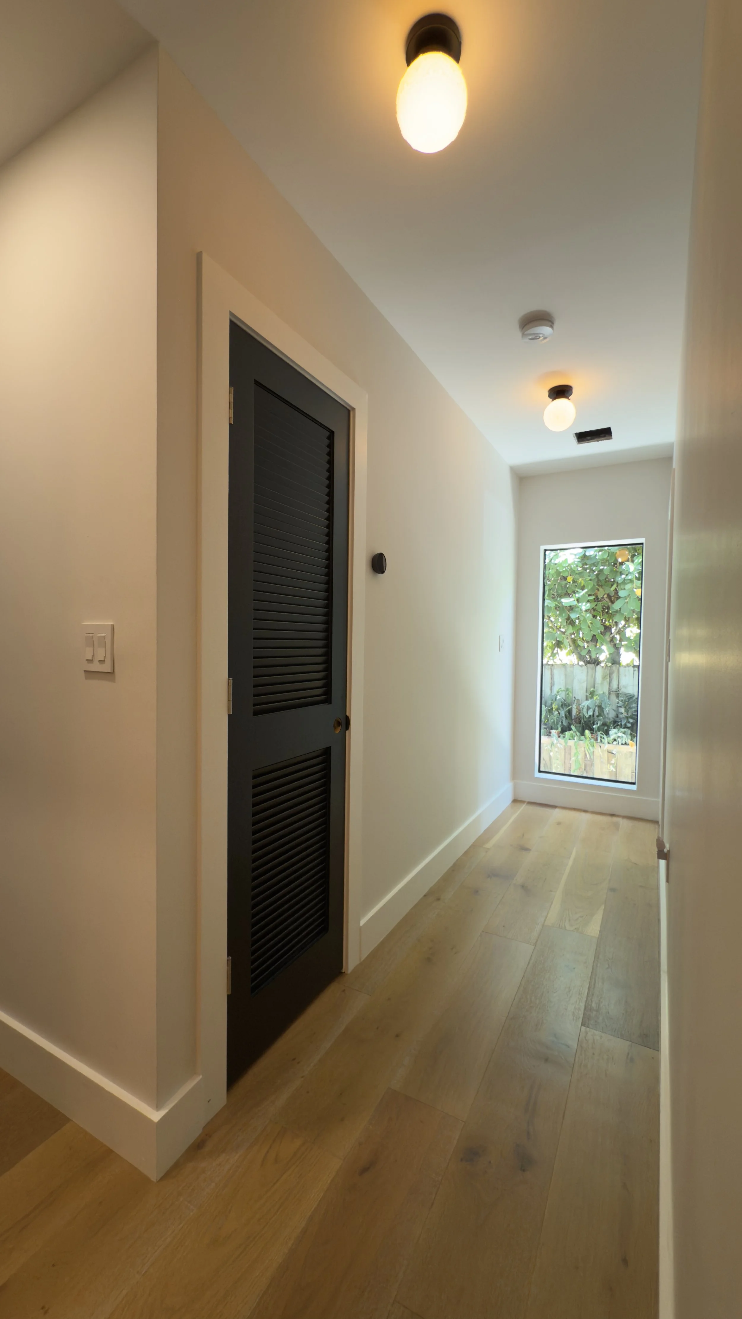 A narrow hallway with wooden flooring, a window showing greenery outside, and two ceiling lights turned on. There is a black louvered door on the left wall.