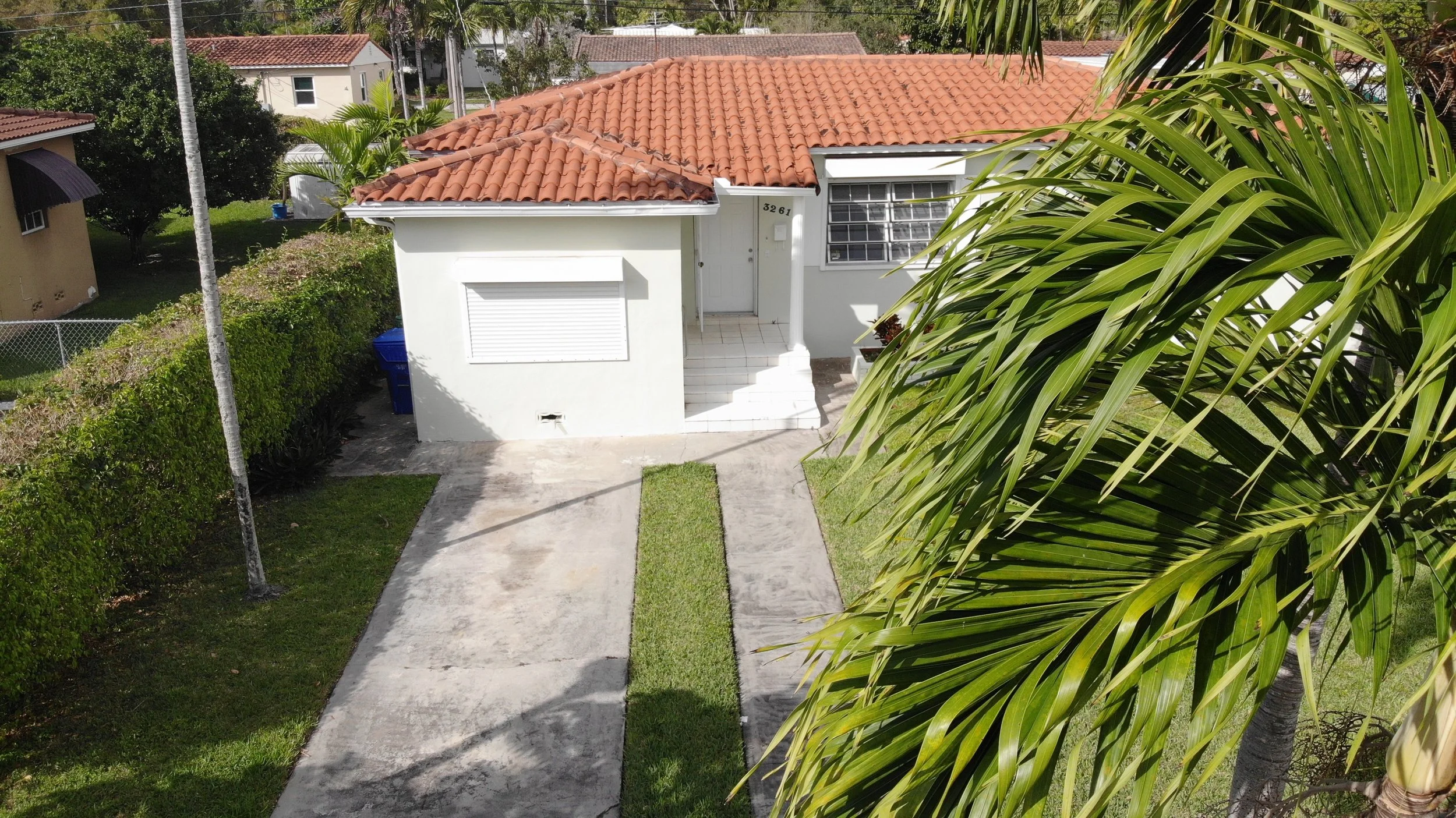 A small white house with a red tile roof, a closed window with shutters, and a small front porch with stairs. The house has the number 3261 near the door, and a white mailbox next to it. There are green shrubs, a concrete driveway, and a large palm t