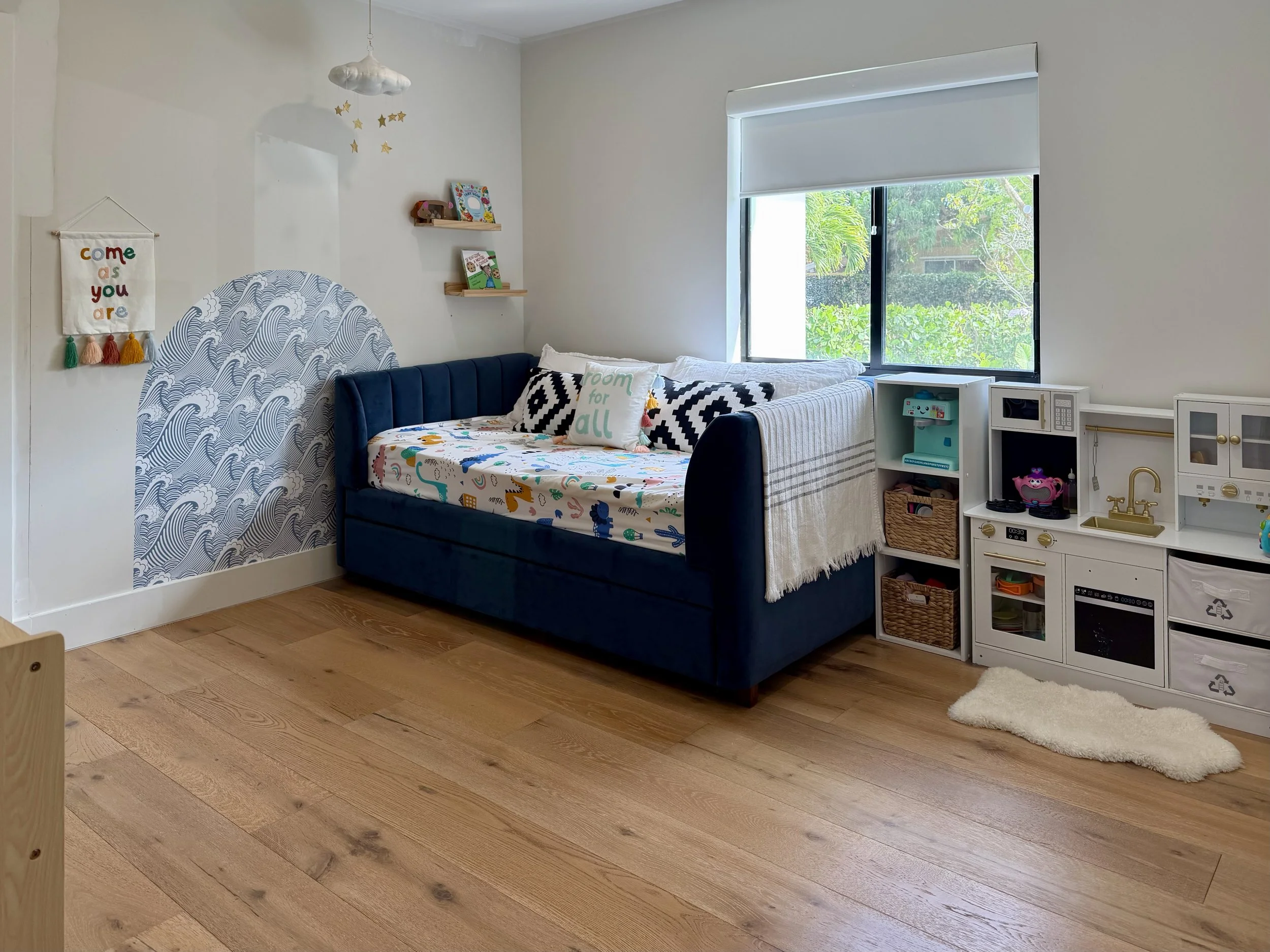A child's bedroom with a navy blue upholstered bed, white wall decorated with a cloud and star chandelier, wall hanging with colorful tassels, wall-mounted shelves with children's books, large window with roller shade, toy kitchen set, plush rug, and