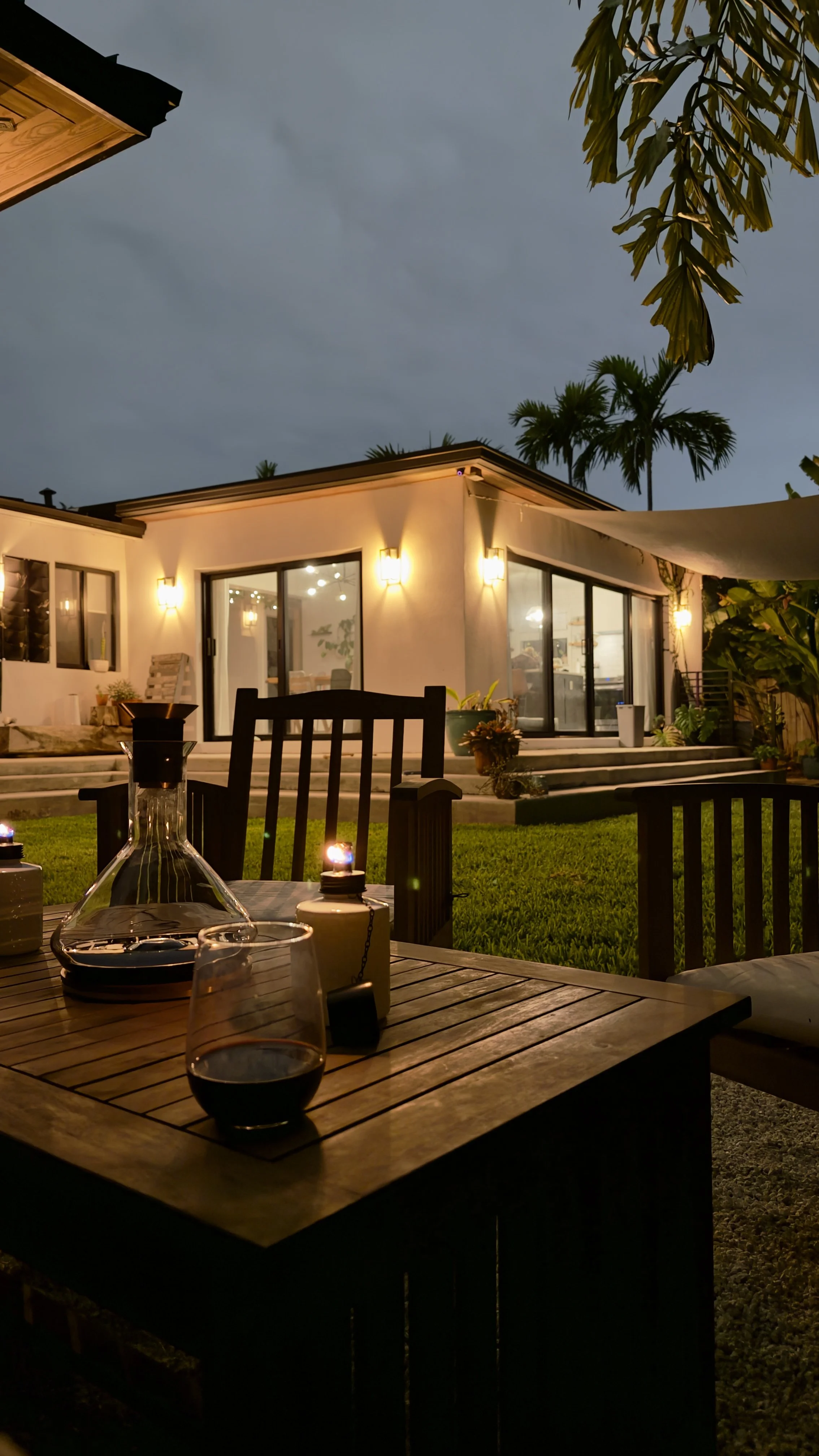 Nighttime outdoor scene of a backyard patio with a wooden table holding a glass of wine, a candle, and a decanter, with a house lit by warm exterior lights in the background, surrounded by lush greenery and palm trees.