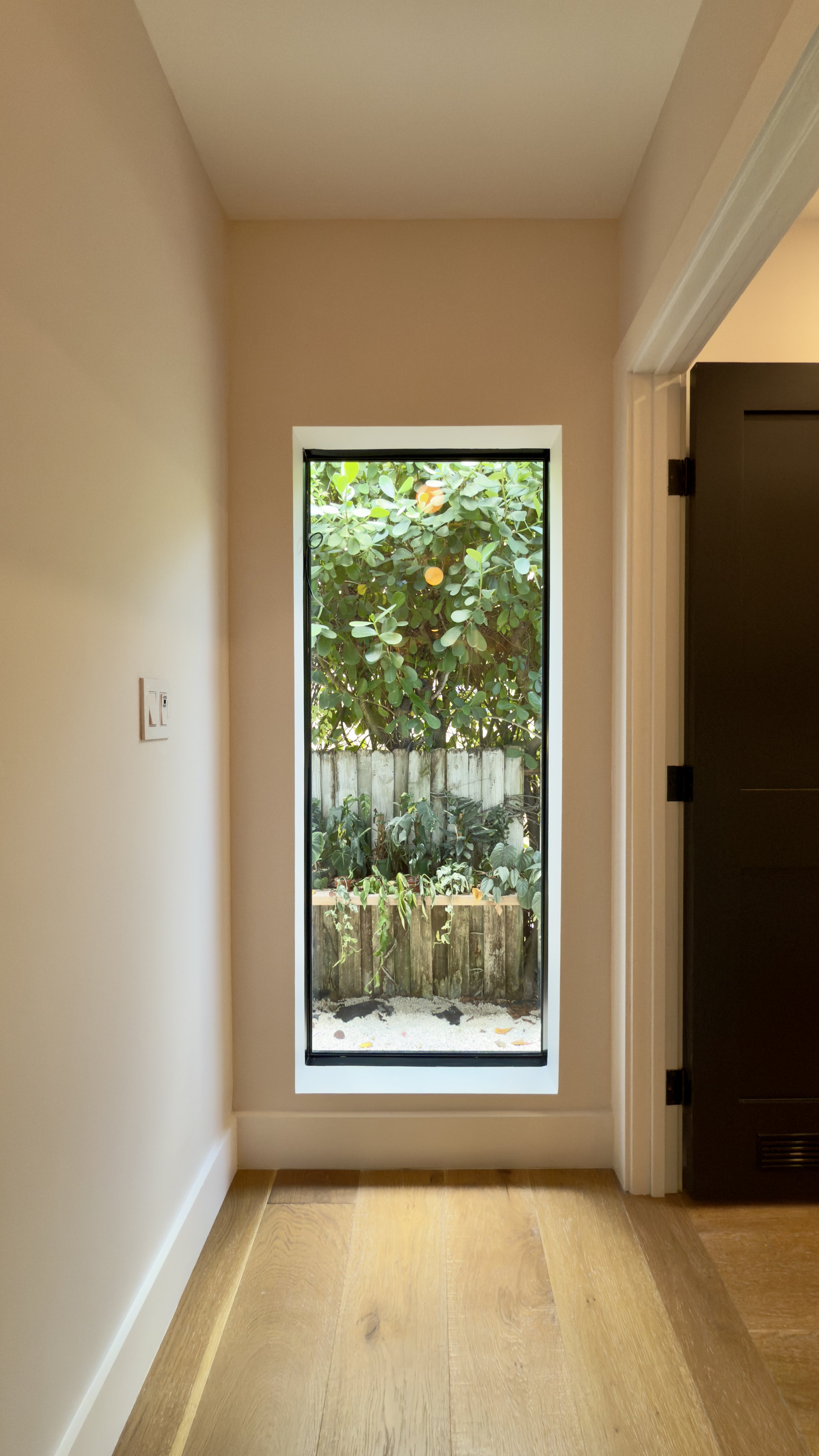 Interior view of a room with hardwood floor, a narrow vertical window showing a garden with green leafy plants and a wooden fence, beige walls, and a black door on the right.