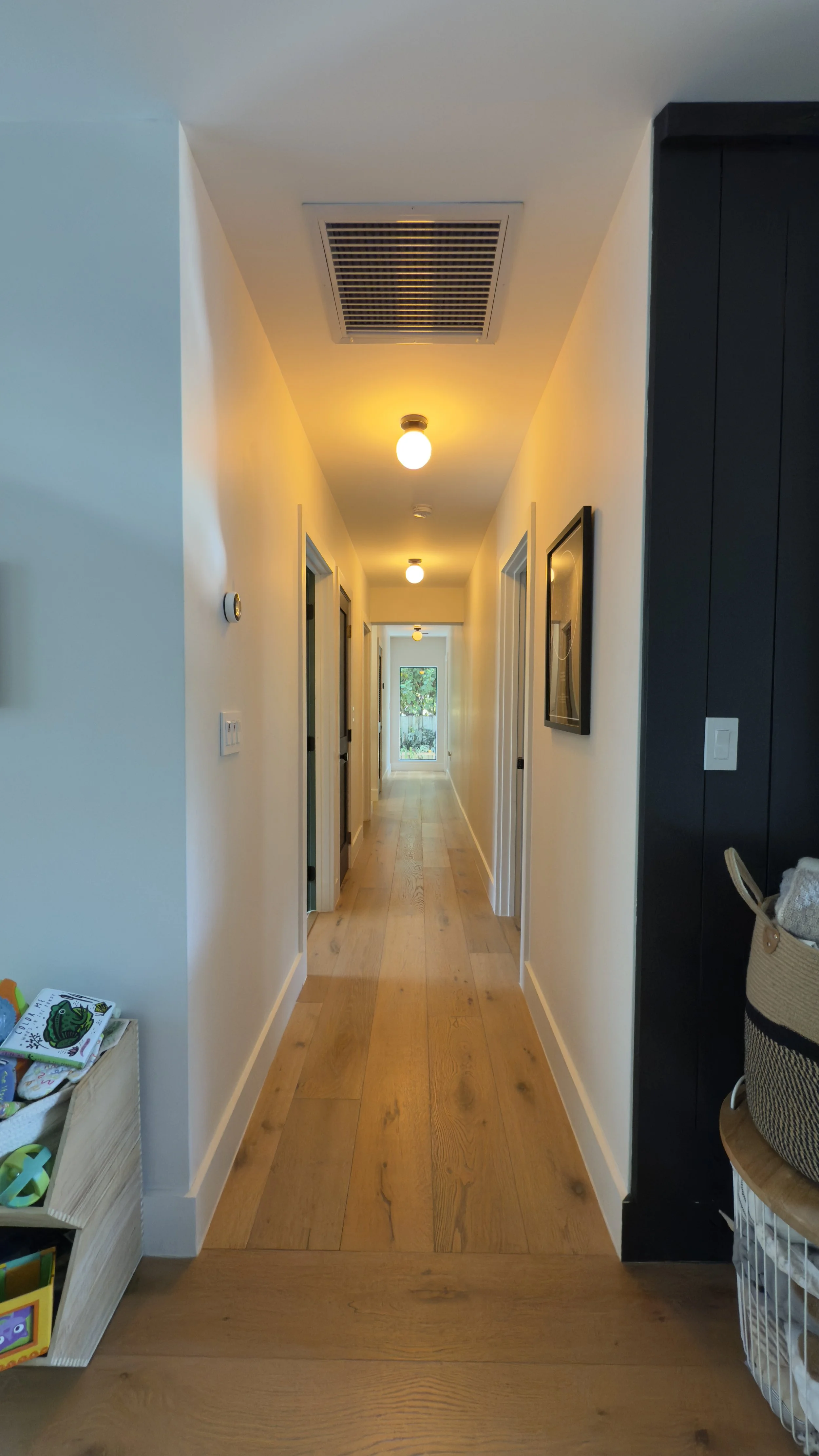 A hallway with wooden flooring, white walls, ceiling lights, and a door at the end with a window revealing greenery outside.