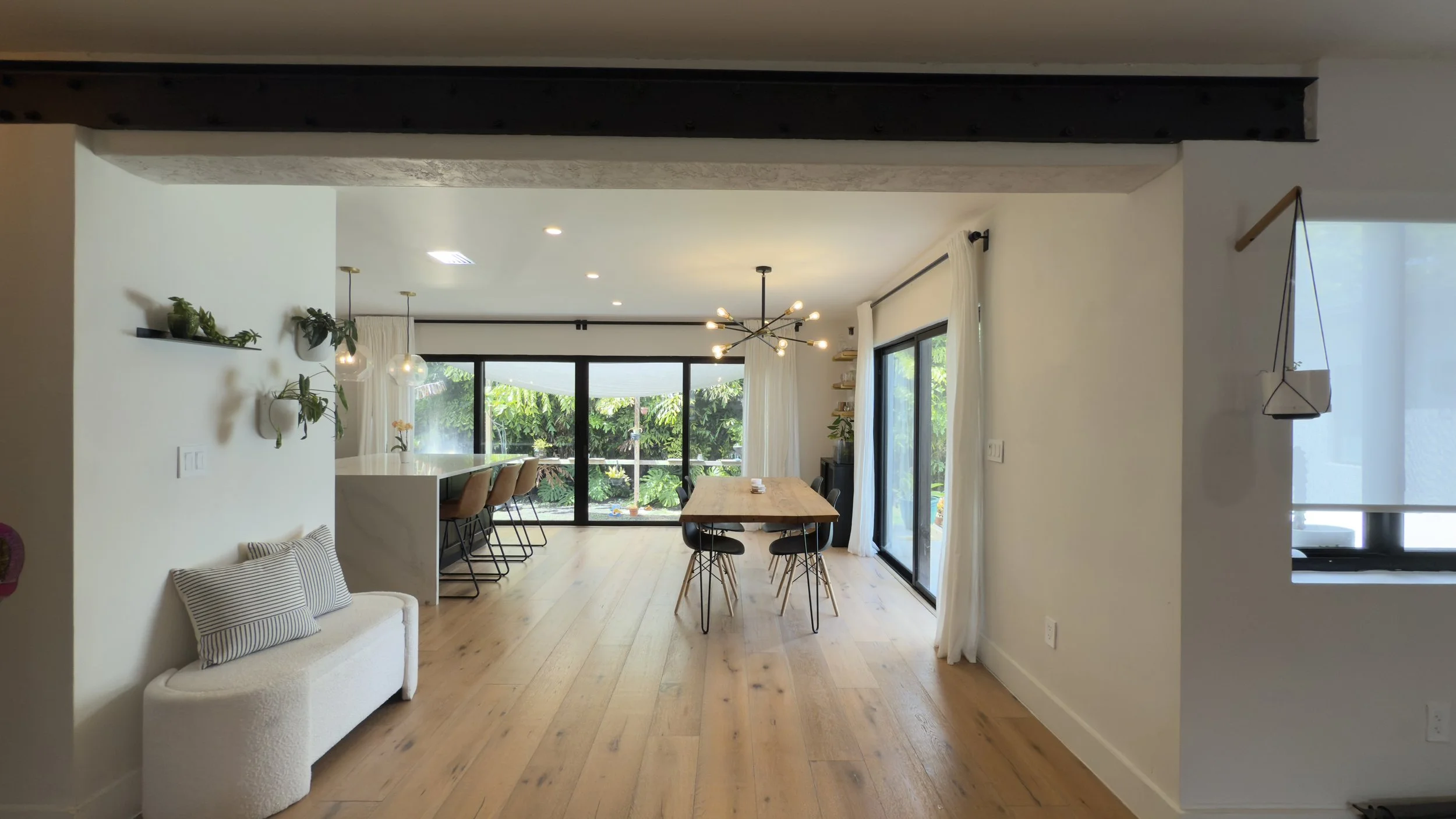 Open-concept dining and living area with wooden floors, a dining table with black chairs, a kitchen island with barstools, and large sliding glass doors leading to a garden.