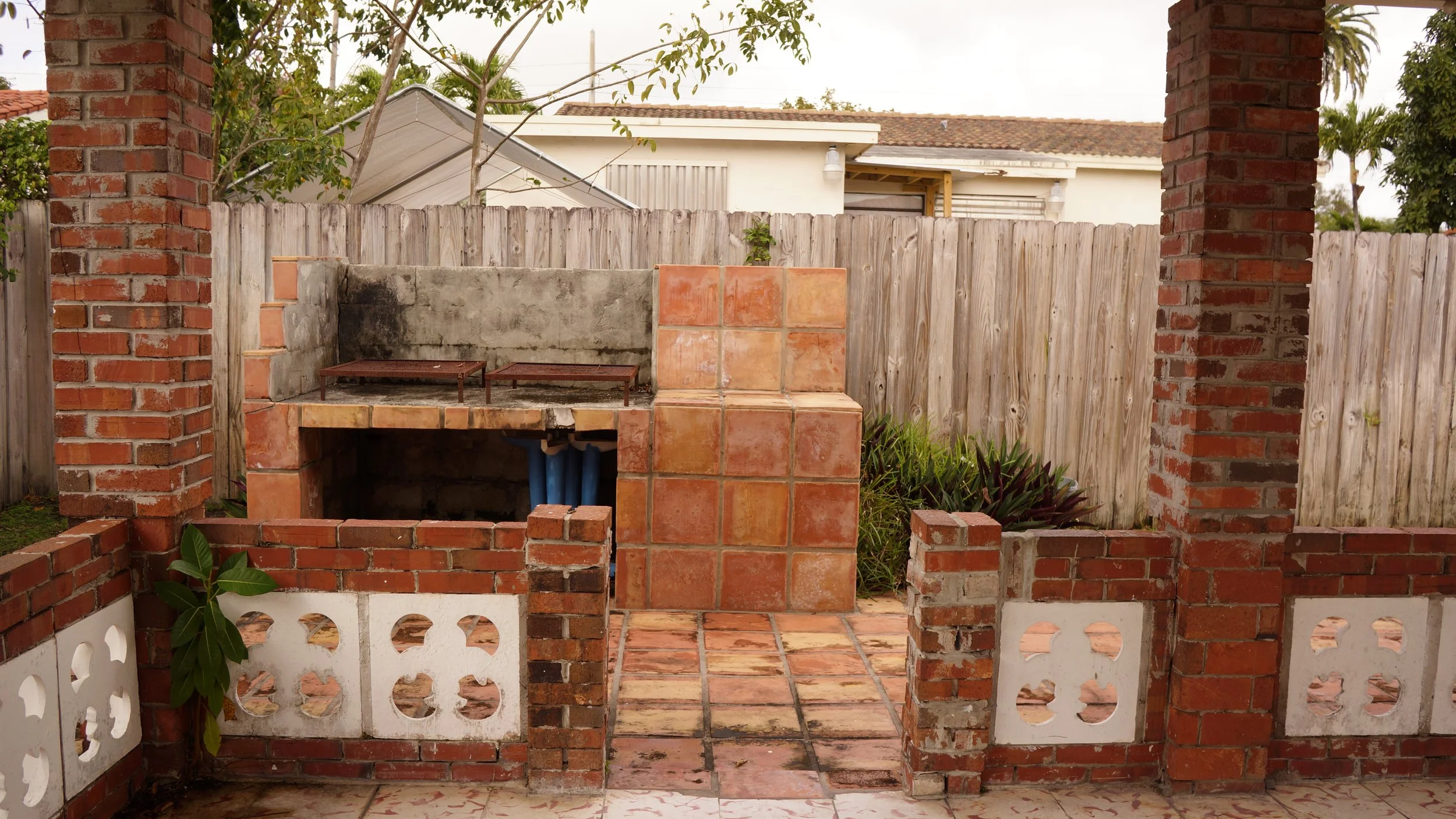 A rustic outdoor brick barbecue area with a fenced backyard, a concrete wall, potted plants, and a tiled patio.