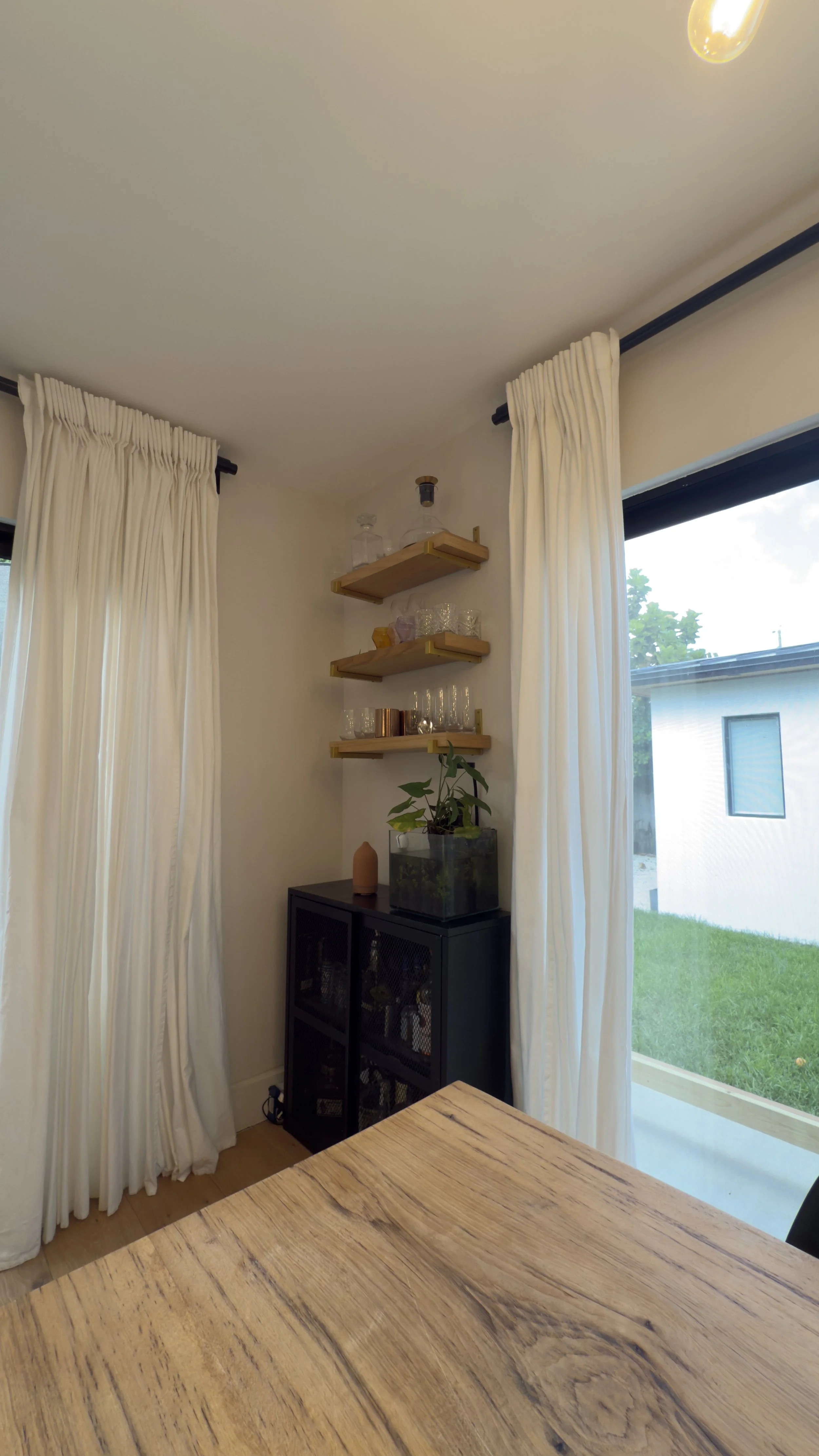 Corner of a room with white curtains, a black cabinet, wooden shelves with glassware, a potted plant, and a window with a view of a grassy yard and a neighboring house.