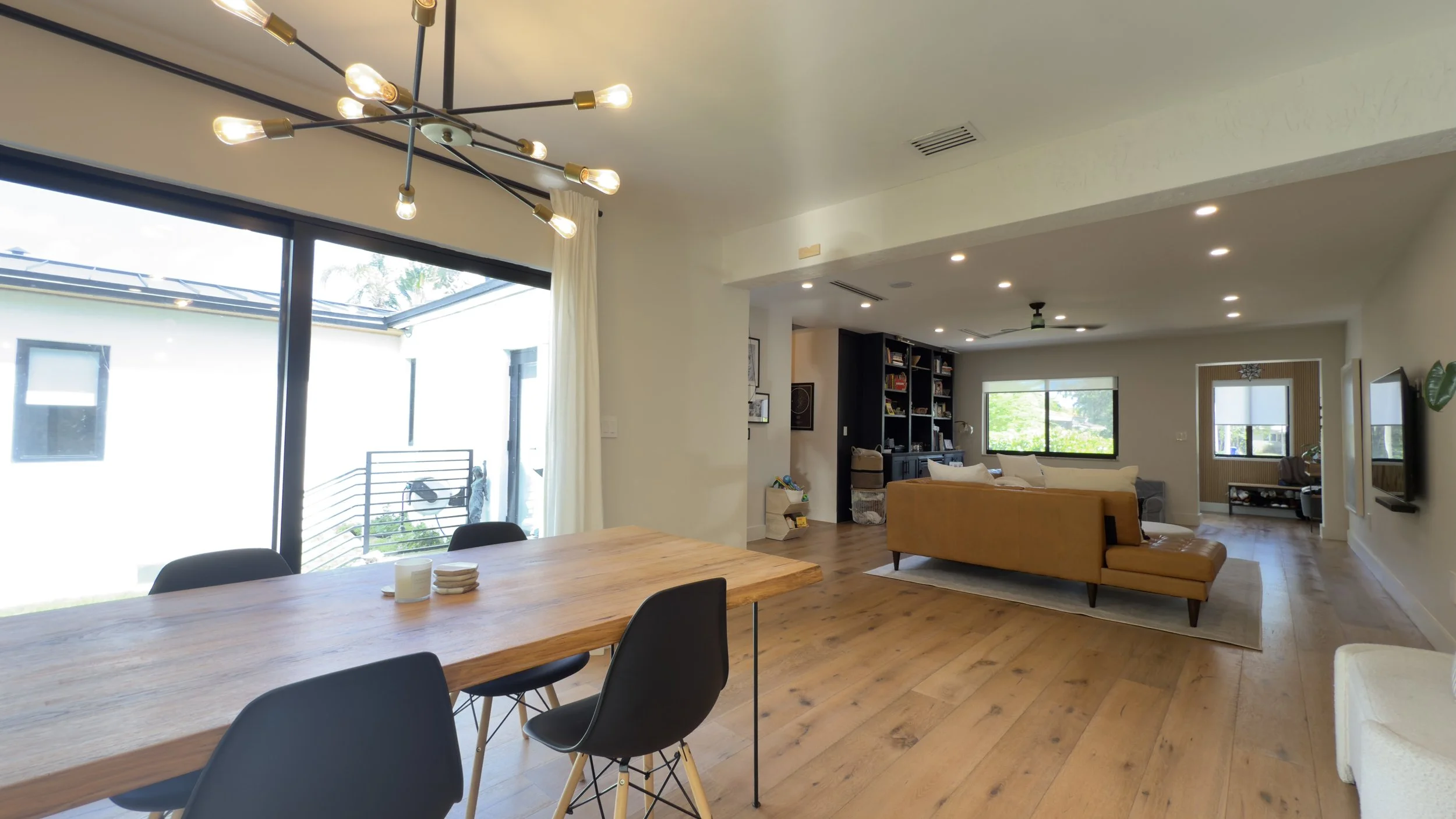 Open-concept living and dining area with wooden flooring, a wooden dining table with black chairs, a yellow sofa, a black bookshelf, walls with framed art, large windows, and modern lighting fixtures.