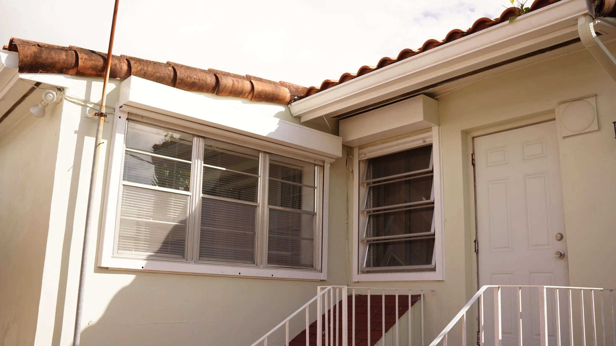 The exterior of a small house with two large windows, a white door, and a metal staircase with white railings. The house has a red tiled roof and a gutter system.