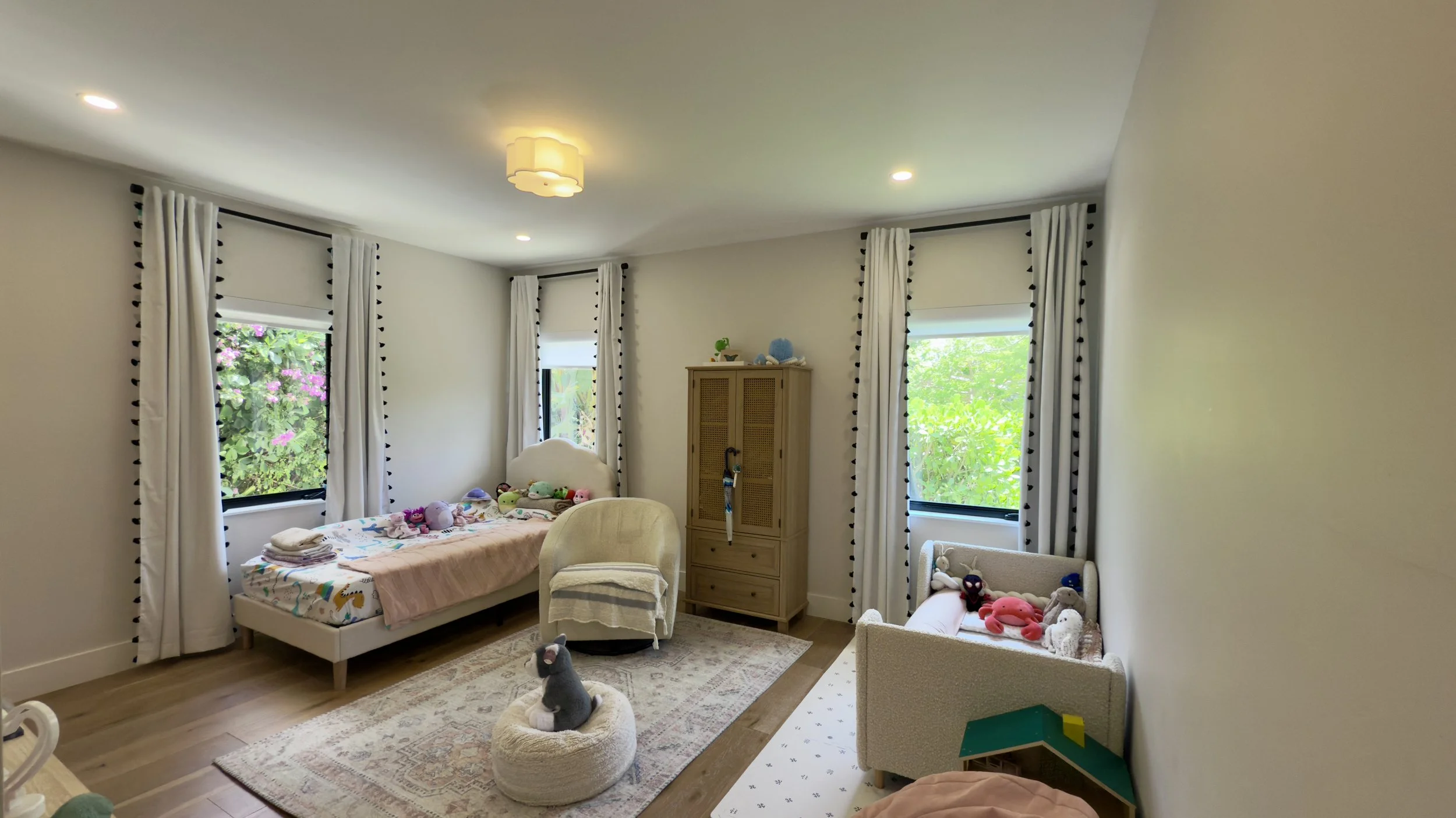 A children's bedroom with three windows, white curtains with black pom-pom trim, a single bed with plush toys, a small armchair, a wooden cabinet, a small sofa with stuffed animals, a beige rug, and a plush cat on a round cushion.
