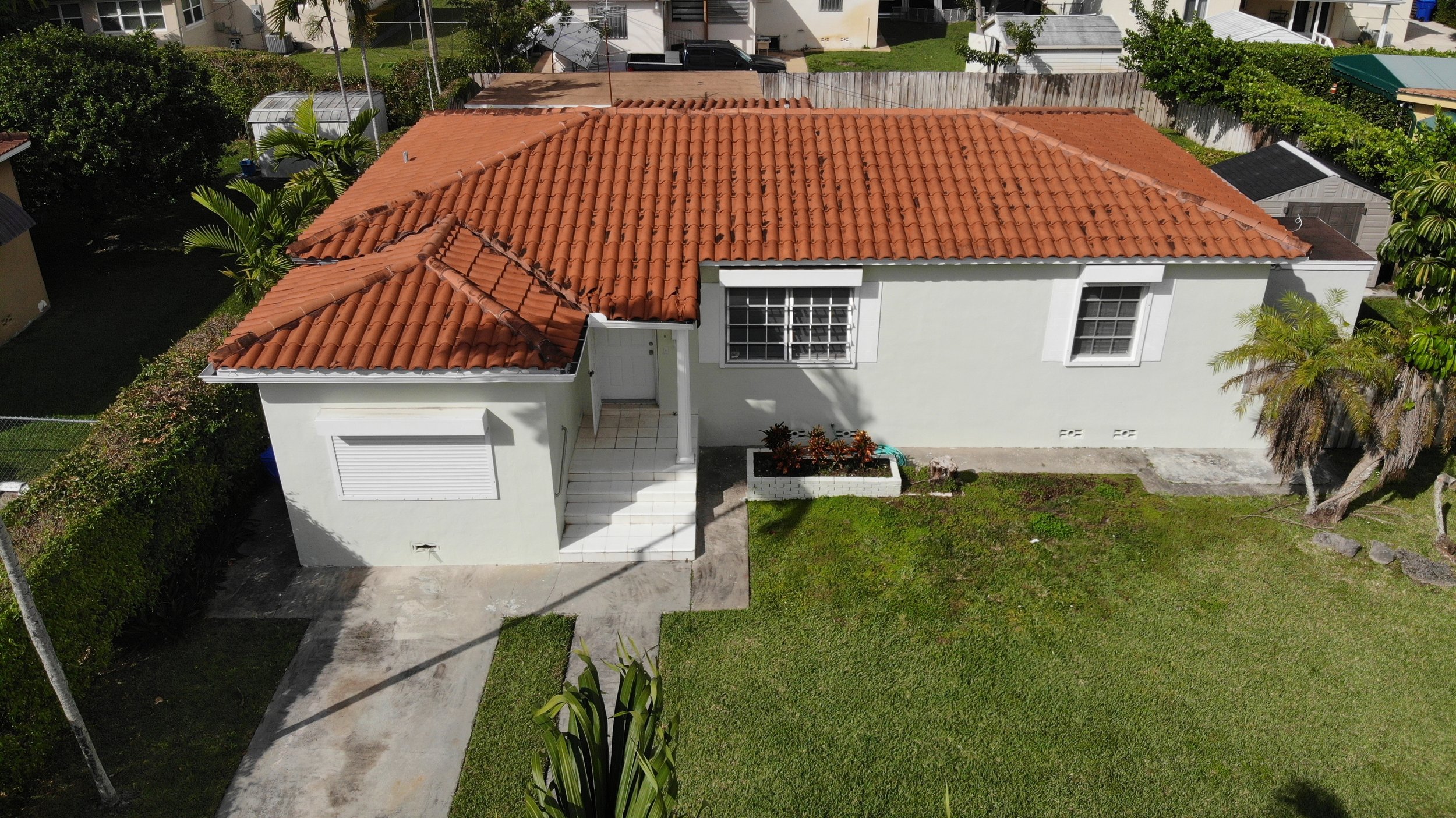 An aerial view of a single-story house with a red tile roof, white exterior walls, and a front combination of tiled steps leading to the entrance and a small garden bed with plants. The house is surrounded by a lawn and has neighboring houses and tre