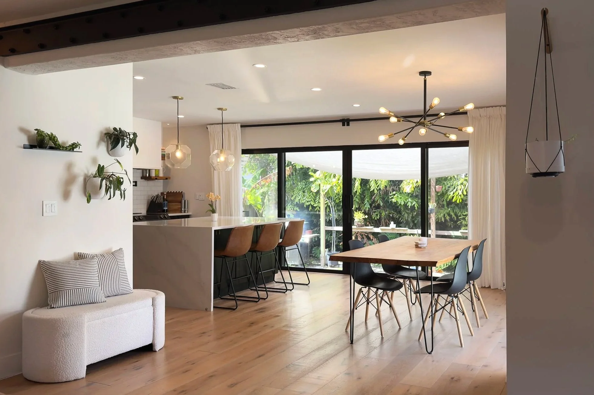 Modern open-concept living and dining area with sliding glass doors leading to a lush garden, featuring a dining table with six chairs, a white kitchen island with three brown bar stools, decorative hanging lights, and a small white couch with stripe