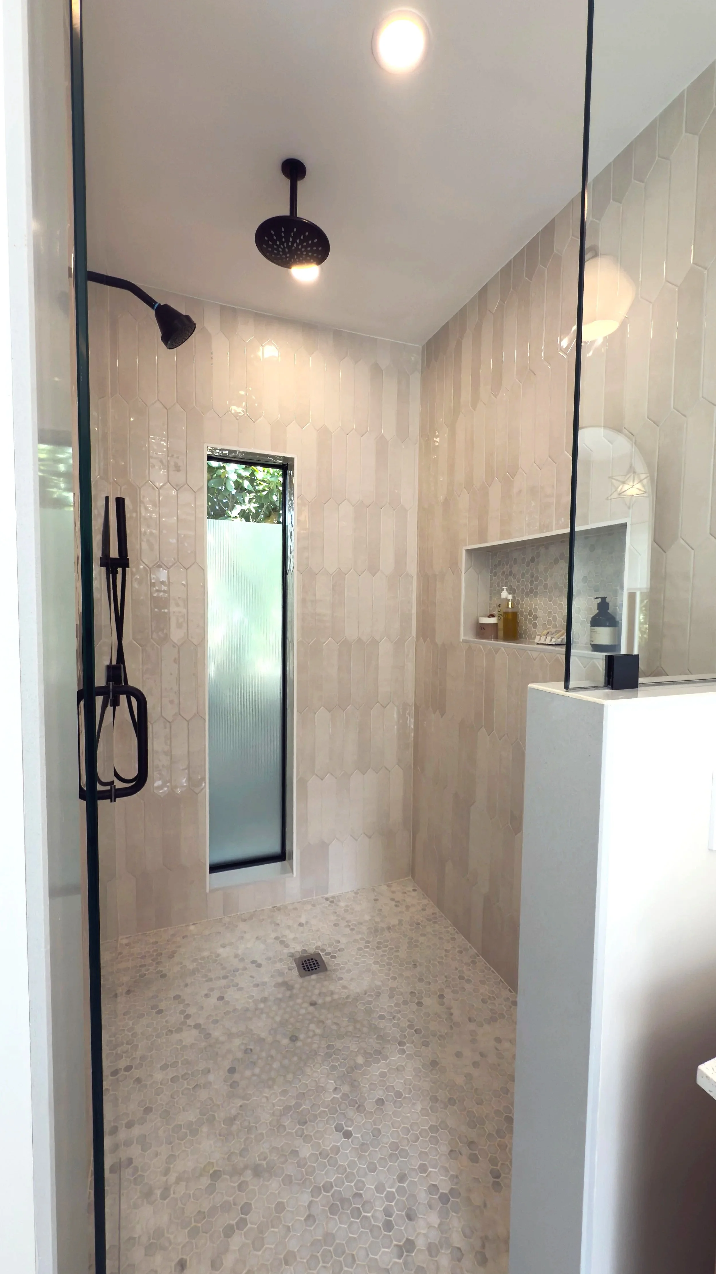 Modern walk-in shower with hexagonal tile walls and pebble tile floor, featuring a niche with toiletries and a frosted window.