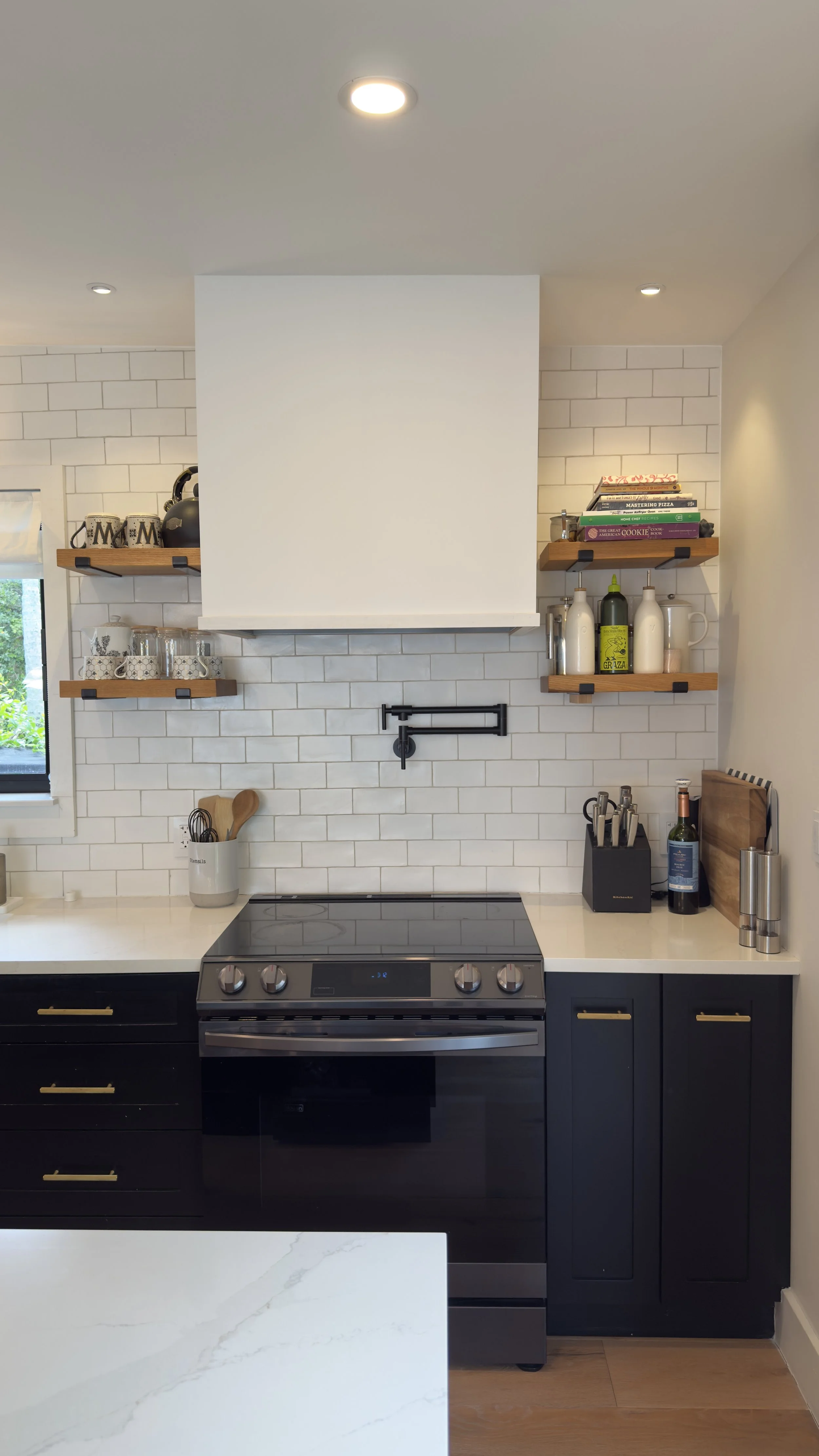 Modern kitchen with black cabinets, white countertop, white subway tile walls, and open wooden shelves with dishes, cookbooks, and condiments.
